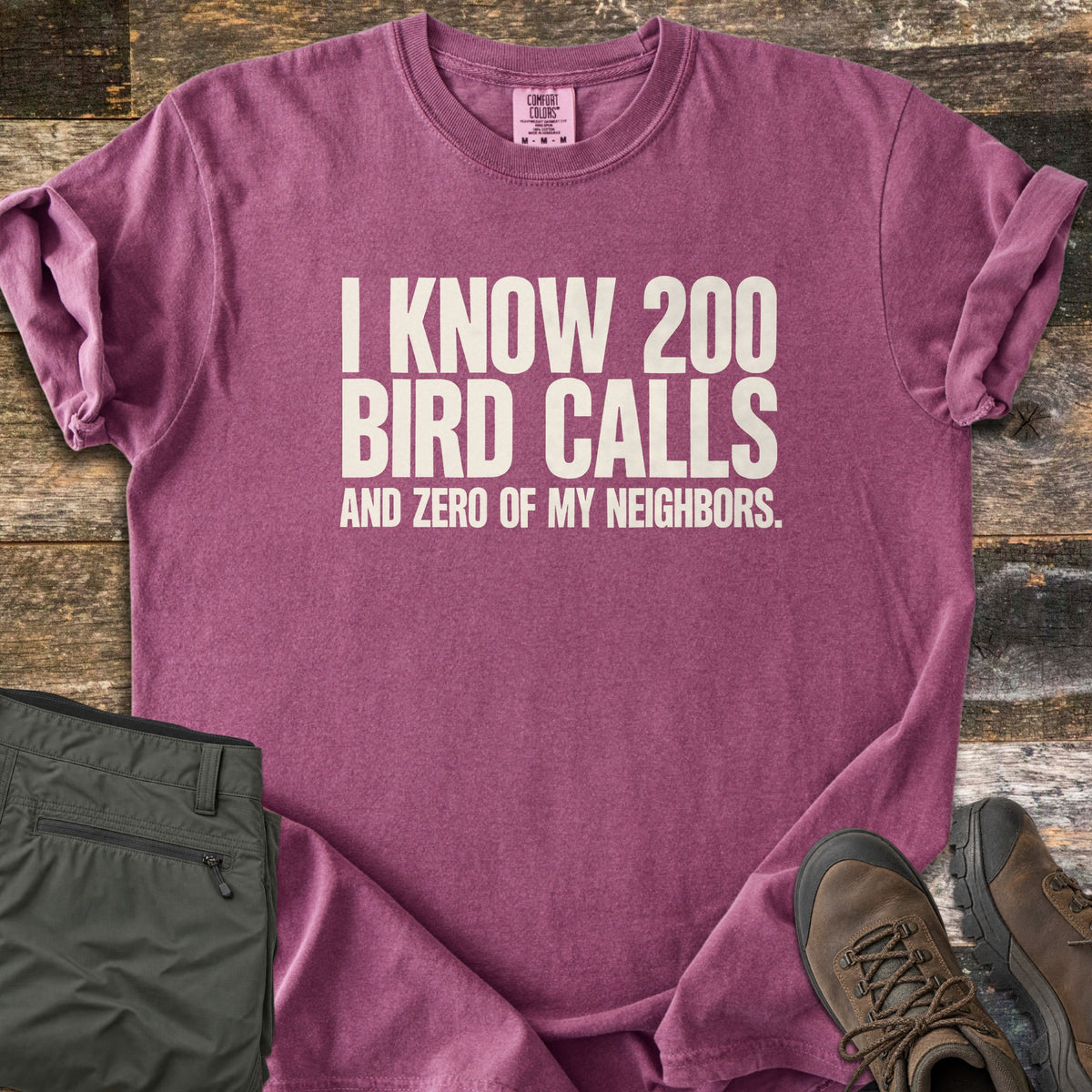 I Know 200 Bird Calls and Zero of My Neighbors Comfort Colors T-shirt in Berry, laid flat. Bold off-white capital letters read I KNOW 200 BIRD CALLS AND ZERO OF MY NEIGHBORS. The shirt has rolled sleeves and lies on weathered wood boards beside folded olive hiking pants and brown hiking boots.