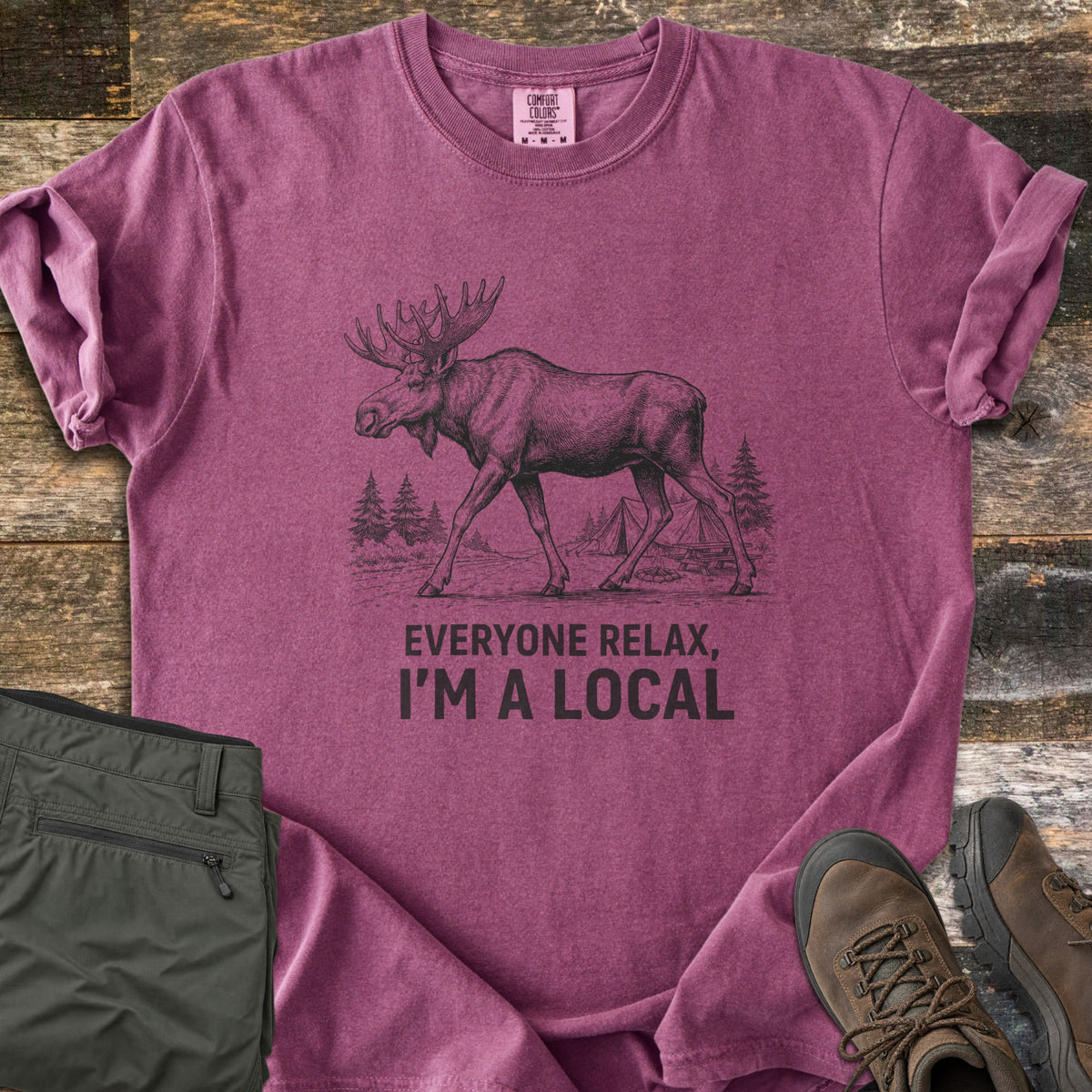 Everyone Relax I'm A Local Comfort Colors T-shirt in Berry, laid flat. A sketched moose walks past pine trees and tents at a campsite above the words Everyone Relax, I'm A Local. Sleeves are rolled on a wood floor beside folded green pants and brown hiking boots.