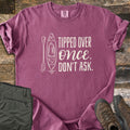Tipped Over Once. Don't Ask. Comfort Colors T-shirt in Berry, laid flat. A white line drawing of a kayak and paddle sits beside the words Tipped Over Once. Don't Ask., with Once in script. Sleeves rolled, on weathered wood with folded olive hiking pants and brown hiking boots.