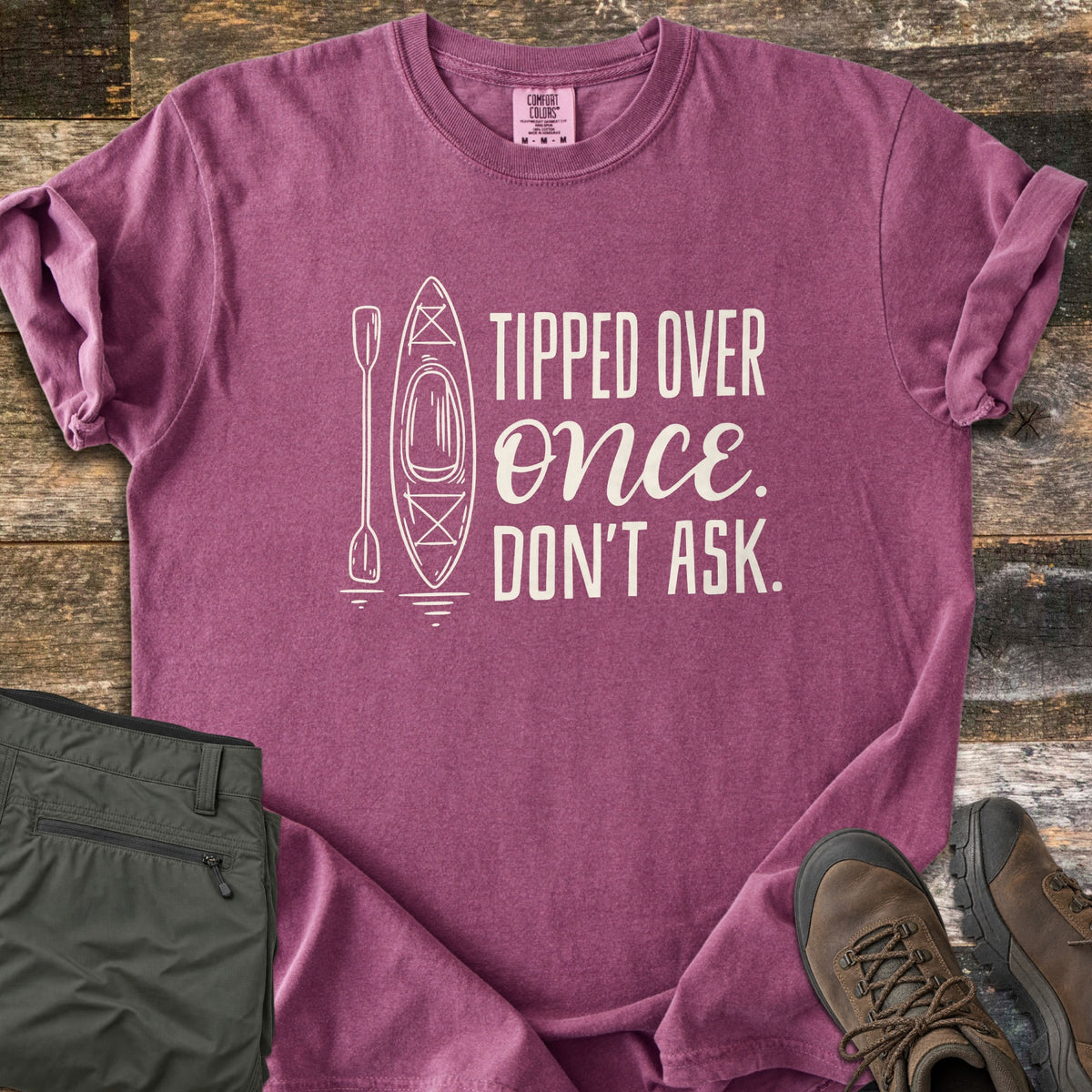 Tipped Over Once. Don't Ask. Comfort Colors T-shirt in Berry, laid flat. A white line drawing of a kayak and paddle sits beside the words Tipped Over Once. Don't Ask., with Once in script. Sleeves rolled, on weathered wood with folded olive hiking pants and brown hiking boots.