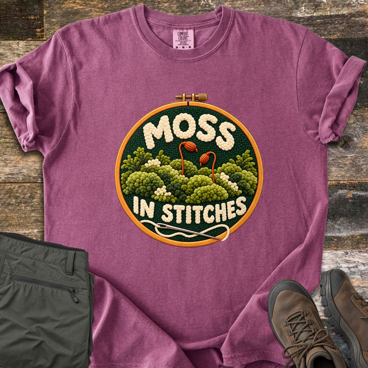 Moss in Stitches Comfort Colors T-shirt in Berry, laid flat. An embroidery hoop holds stitched green moss with two orange spore capsules, the word Moss above and In Stitches below, plus a needle and thread. Folded olive pants and brown hiking boots lie beside it on weathered wood.