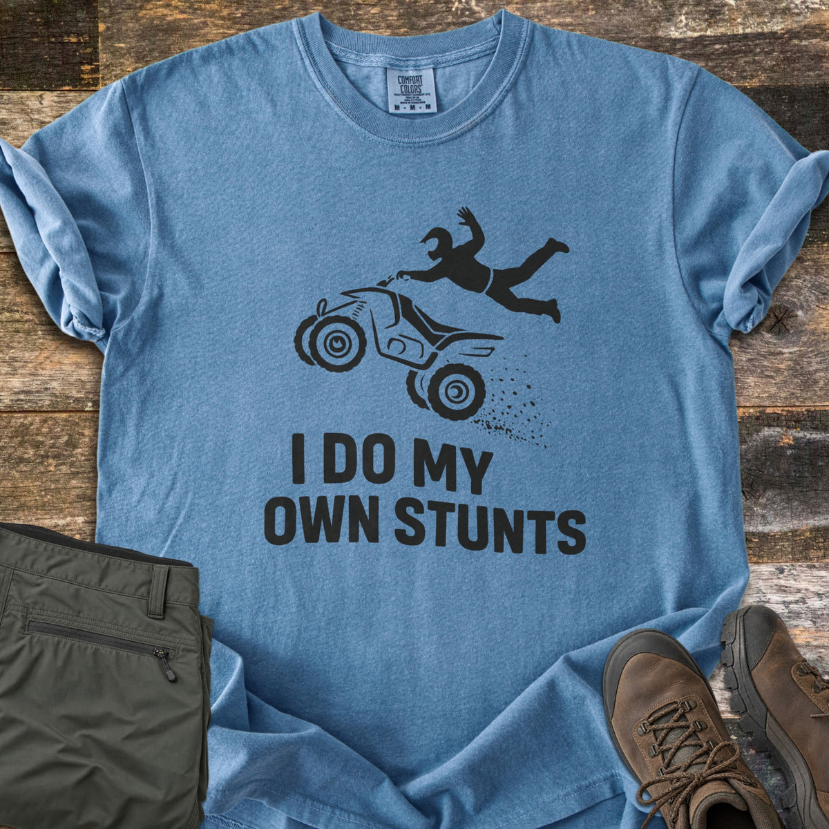 ATV Stunts Comfort Colors T-shirt in Blue Jean, laid flat. A black silhouette of a helmeted rider flying off a wheelie-popping ATV kicking up dirt, above the words I Do My Own Stunts. Olive cargo pants and brown hiking boots sit beside it on weathered wood.