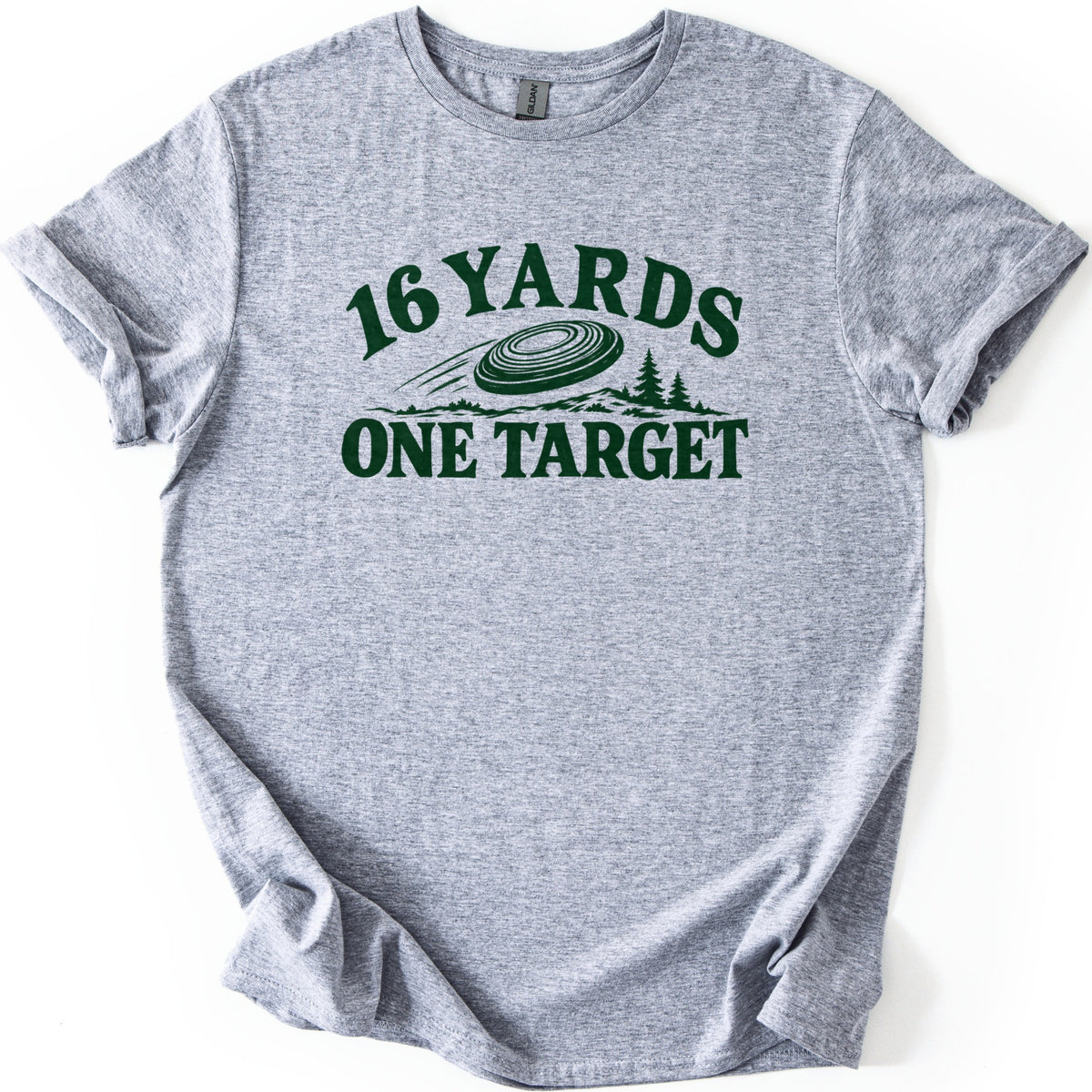 16 Yards One Target Comfort Colors T-shirt in Sport Grey, laid flat. Dark green artwork shows a clay target flying over hills and pine trees, with 16 YARDS curved above and ONE TARGET below. The shirt has rolled sleeves and lies on a white background.