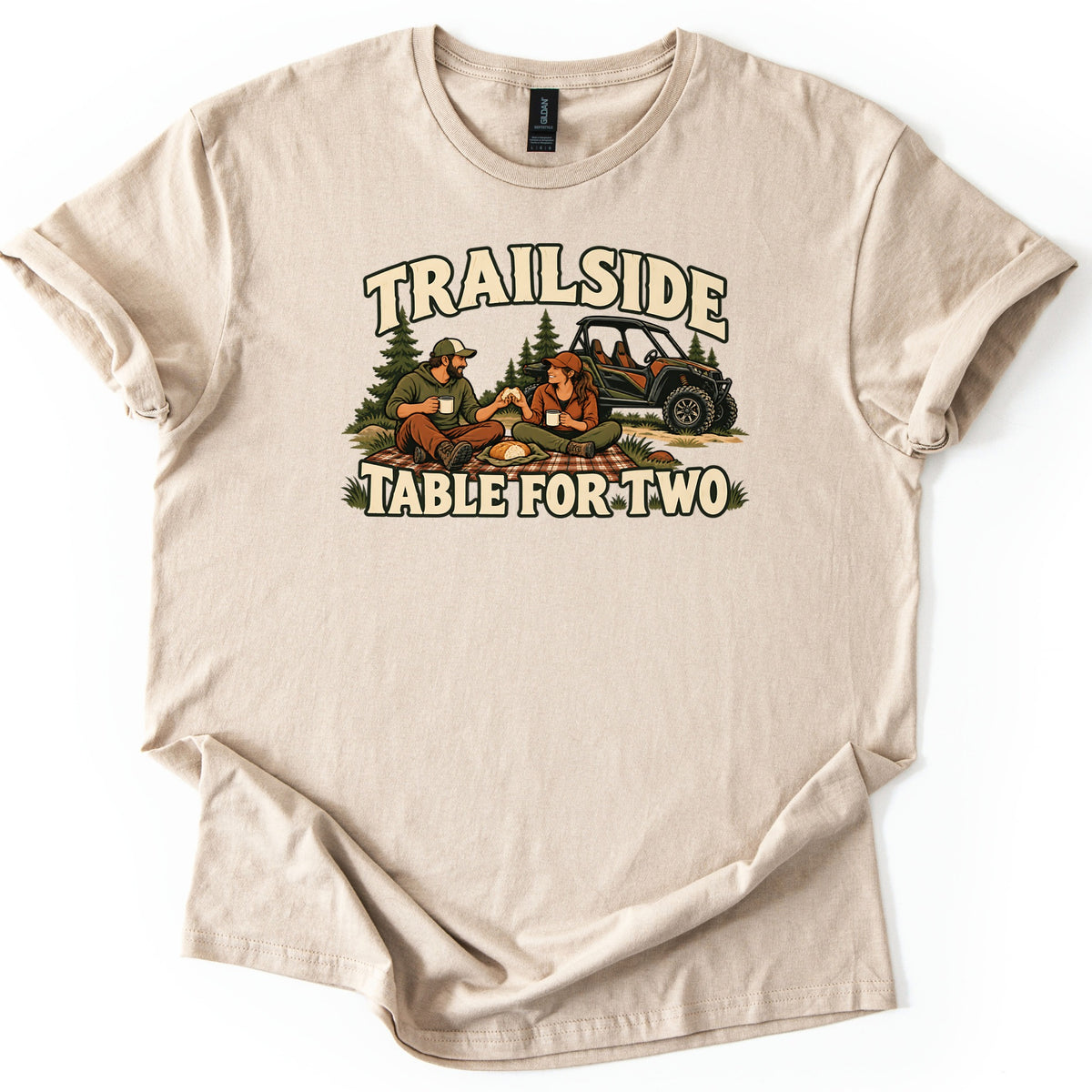 Trailside Table for Two Comfort Colors T-shirt in Sand, laid flat. Trailside arches over a couple in caps sharing a picnic on a plaid blanket with mugs and bread, pine trees and an off-road side-by-side behind, with Table for Two below. Cuffed sleeves, on white.