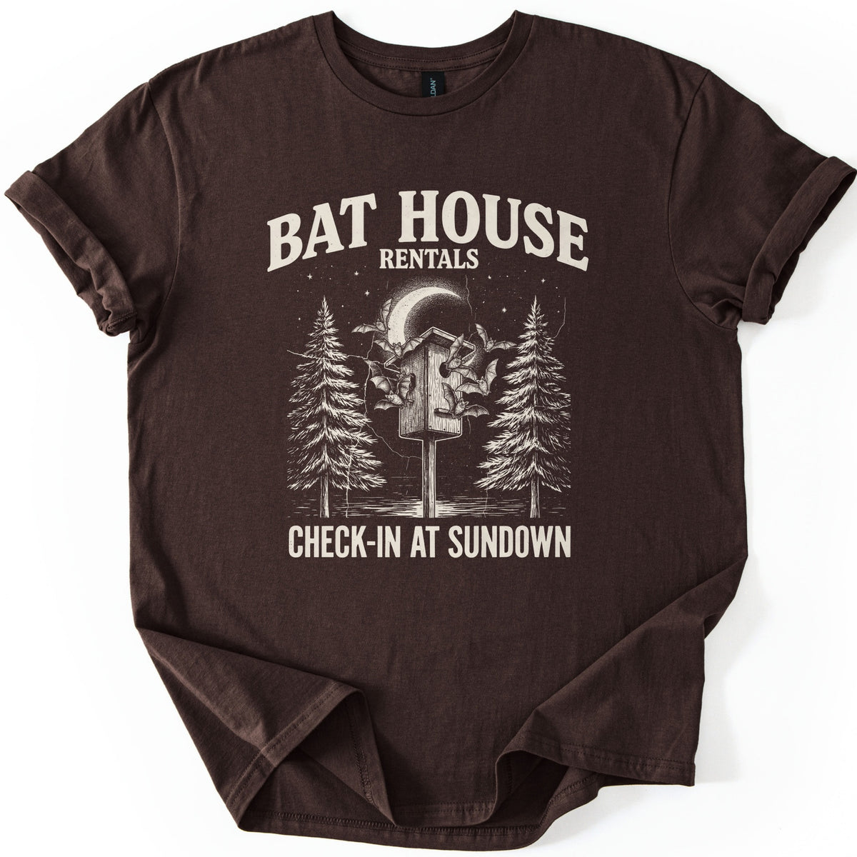 Bat House Rentals Comfort Colors T-shirt in Dark Chocolate, laid flat. Cream text reads Bat House Rentals above a drawing of bats around a wooden bat house on a post under a crescent moon and stars, framed by two pine trees, with Check-In at Sundown below. Sleeves cuffed, on white.