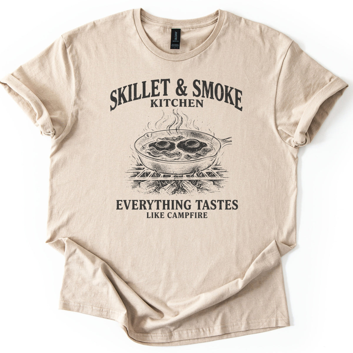 Skillet & Smoke Kitchen Comfort Colors T-shirt in Sand, laid flat. A black print shows a cast iron skillet of fried eggs and bacon steaming on a grate over a campfire, under the words Skillet & Smoke Kitchen and above Everything Tastes Like Campfire. Sleeves cuffed, on white.