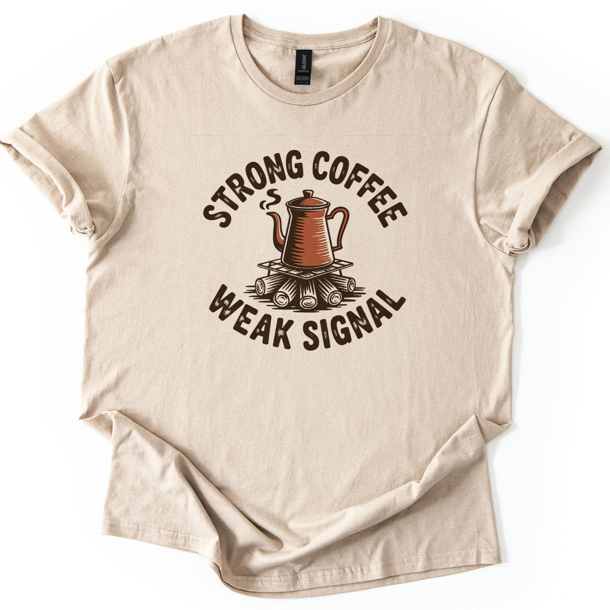 Strong Coffee Weak Signal Comfort Colors T-shirt in Sand, laid flat. A steaming copper coffee pot rests on a grate above campfire logs, with Strong Coffee arched over it and Weak Signal curved beneath in distressed dark brown letters. Cuffed sleeves, white background.