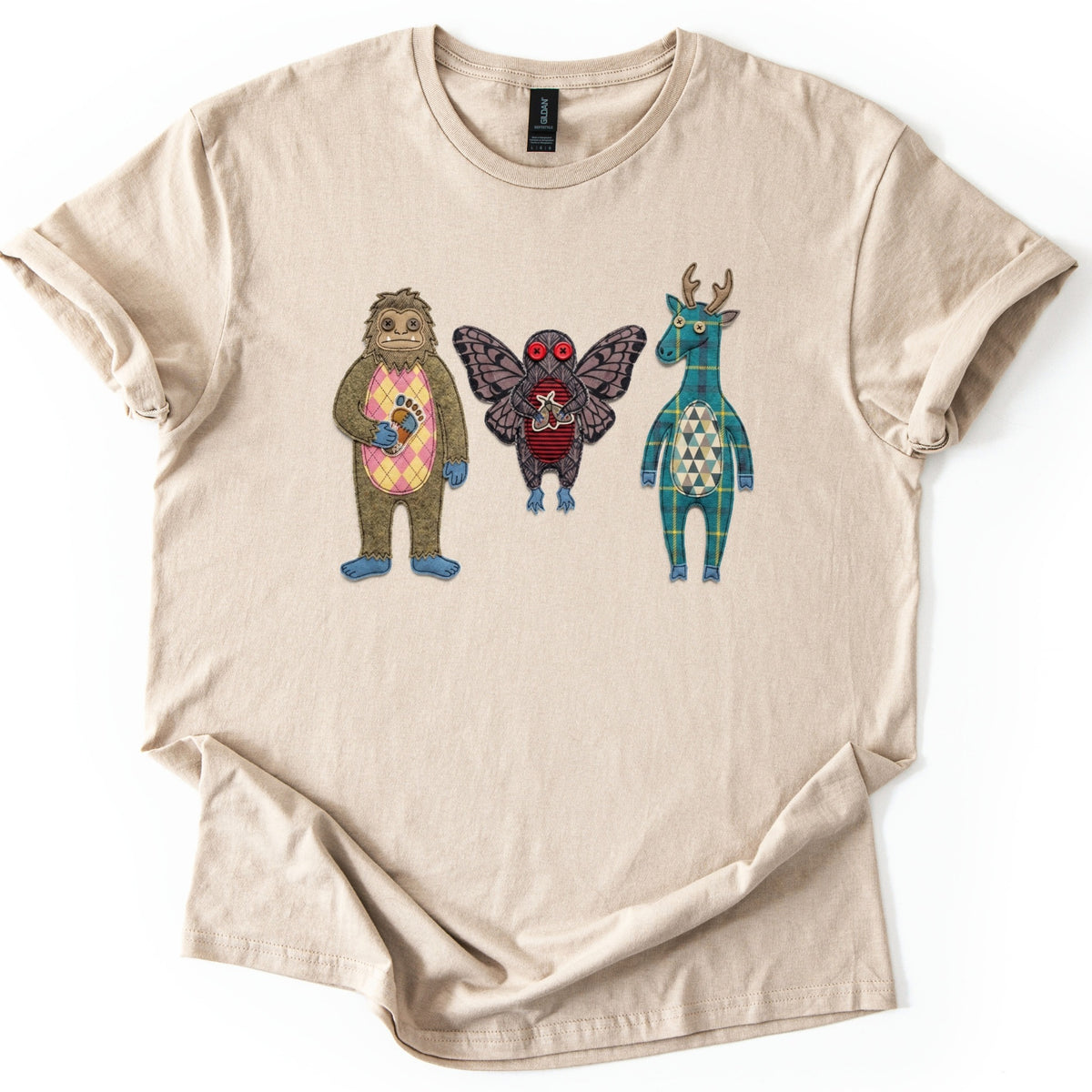 Patchwork Cryptid Trio Comfort Colors T-shirt in Sand, laid flat. Three patchwork-style cryptids with no words: Bigfoot holding a footprint patch, Mothman with moth wings and red button eyes, and an antlered creature in teal plaid. Sleeves cuffed, on white.