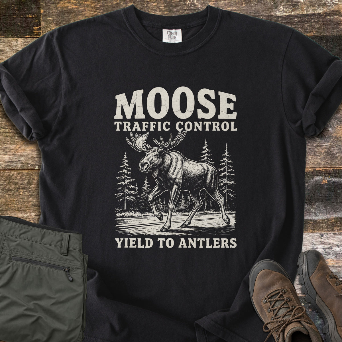 Moose Traffic Control Comfort Colors T-shirt in Black, laid flat. An off-white sketch of a moose walking across a road between pine trees, with MOOSE TRAFFIC CONTROL above and YIELD TO ANTLERS below. Sleeves rolled, on weathered wood with olive pants and brown hiking boots.