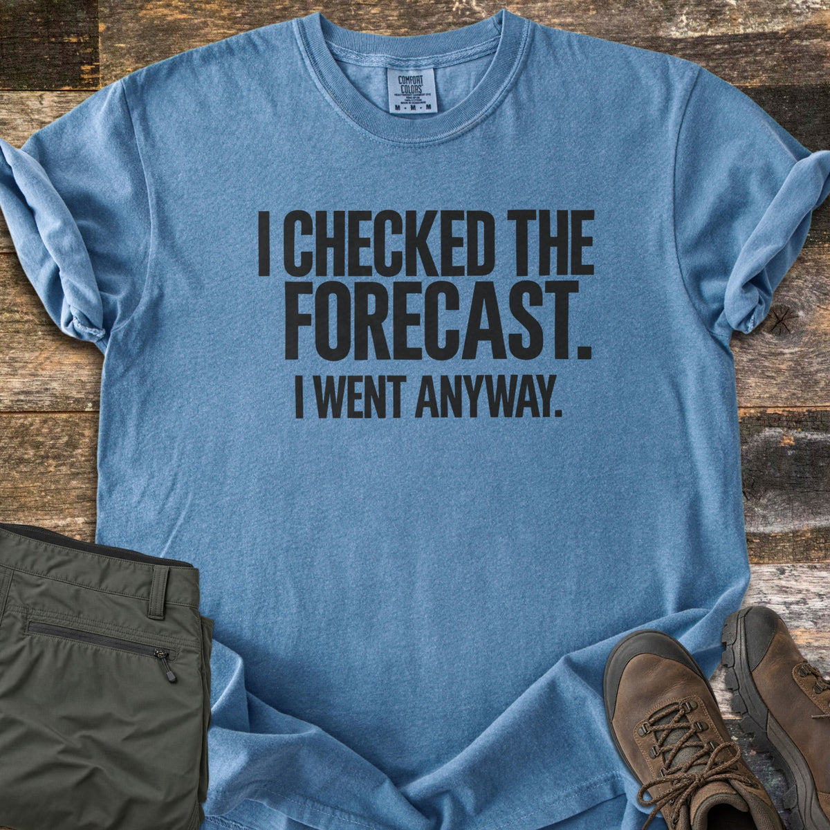 Checked The Forecast Comfort Colors T-shirt in Blue Jean, laid flat. Black block letters read I CHECKED THE FORECAST. I WENT ANYWAY. The sleeves are cuffed, with folded olive hiking pants and brown hiking boots beside it on weathered wood planks.