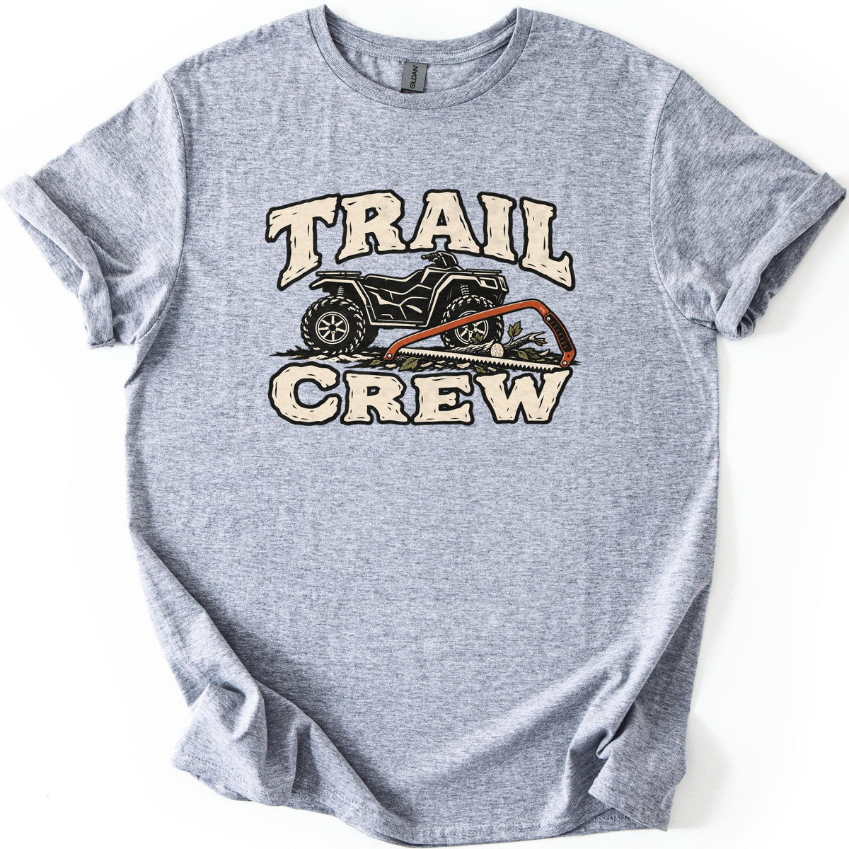 Trail Crew Comfort Colors T-shirt in Sport Grey, laid flat. The words TRAIL and CREW in cream letters with black outlines frame a black ATV and an orange bow saw on a cut branch with leaves. The shirt lies on a white background with rolled sleeves.