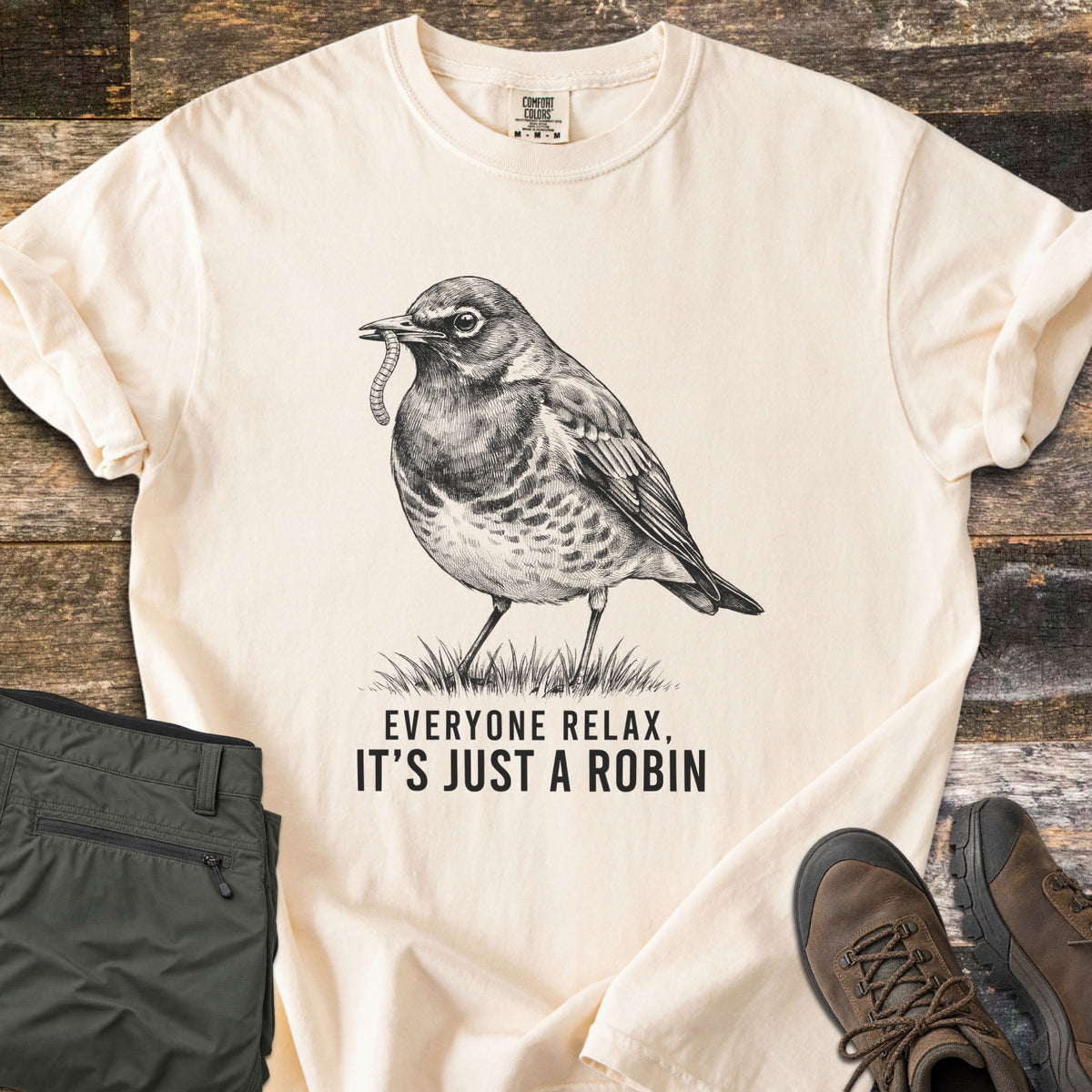 Everyone Relax It's Just A Robin Comfort Colors T-shirt in Ivory, laid flat. A black ink sketch of a robin with a worm in its beak stands on grass above the words Everyone Relax, It's Just A Robin. Olive cargo pants and brown hiking boots lie beside it on weathered wood.