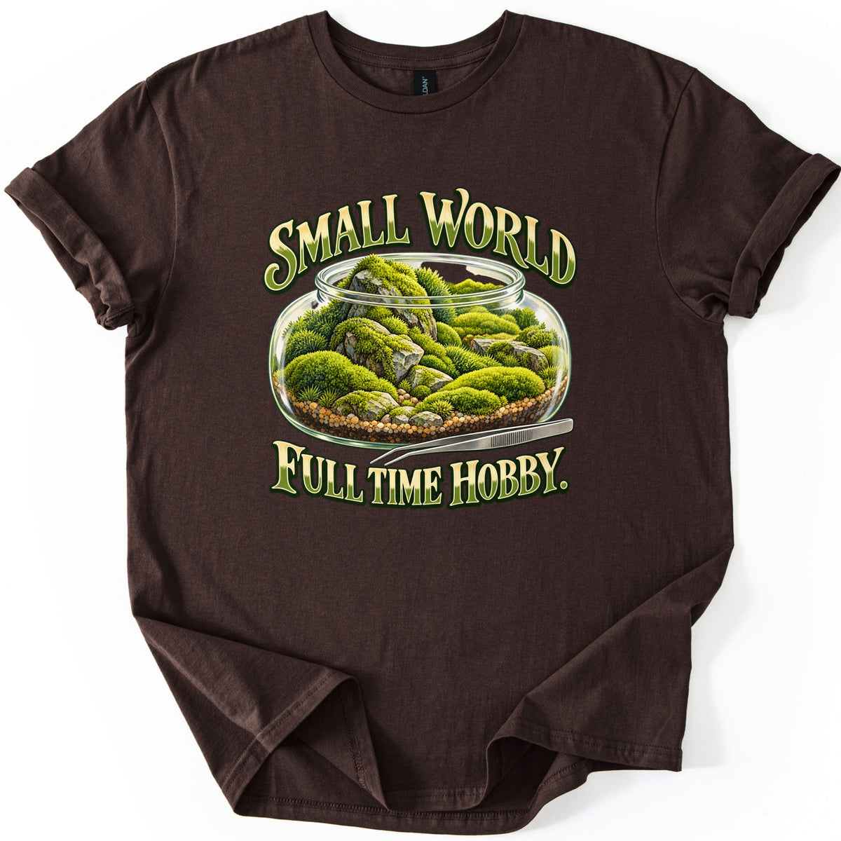 Small World Full Time Hobby Comfort Colors T-shirt in Dark Chocolate, laid flat. A glass terrarium jar of green moss and gray rocks on gravel, with metal tweezers beside it, between the words Small World and Full Time Hobby. in green and gold letters. Sleeves cuffed, on a white background.