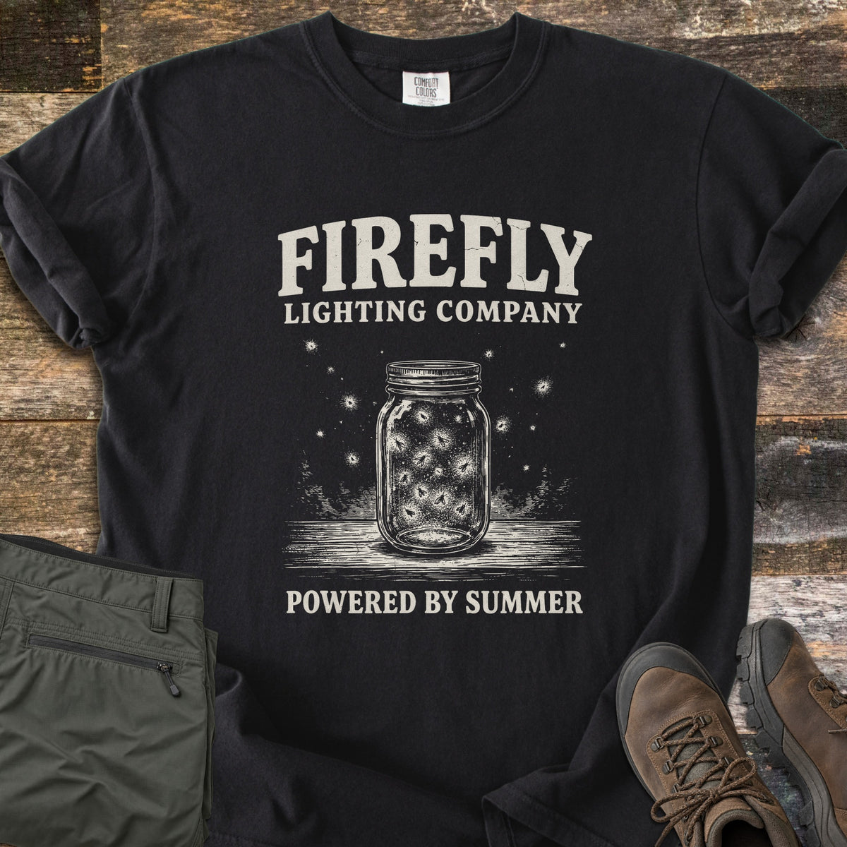 Firefly Lighting Company Comfort Colors T-shirt in Black, laid flat. Cream print of a mason jar full of glowing fireflies on a wooden surface, under the words Firefly Lighting Company and above Powered by Summer. Olive cargo pants and brown hiking boots lie beside it on weathered wood.