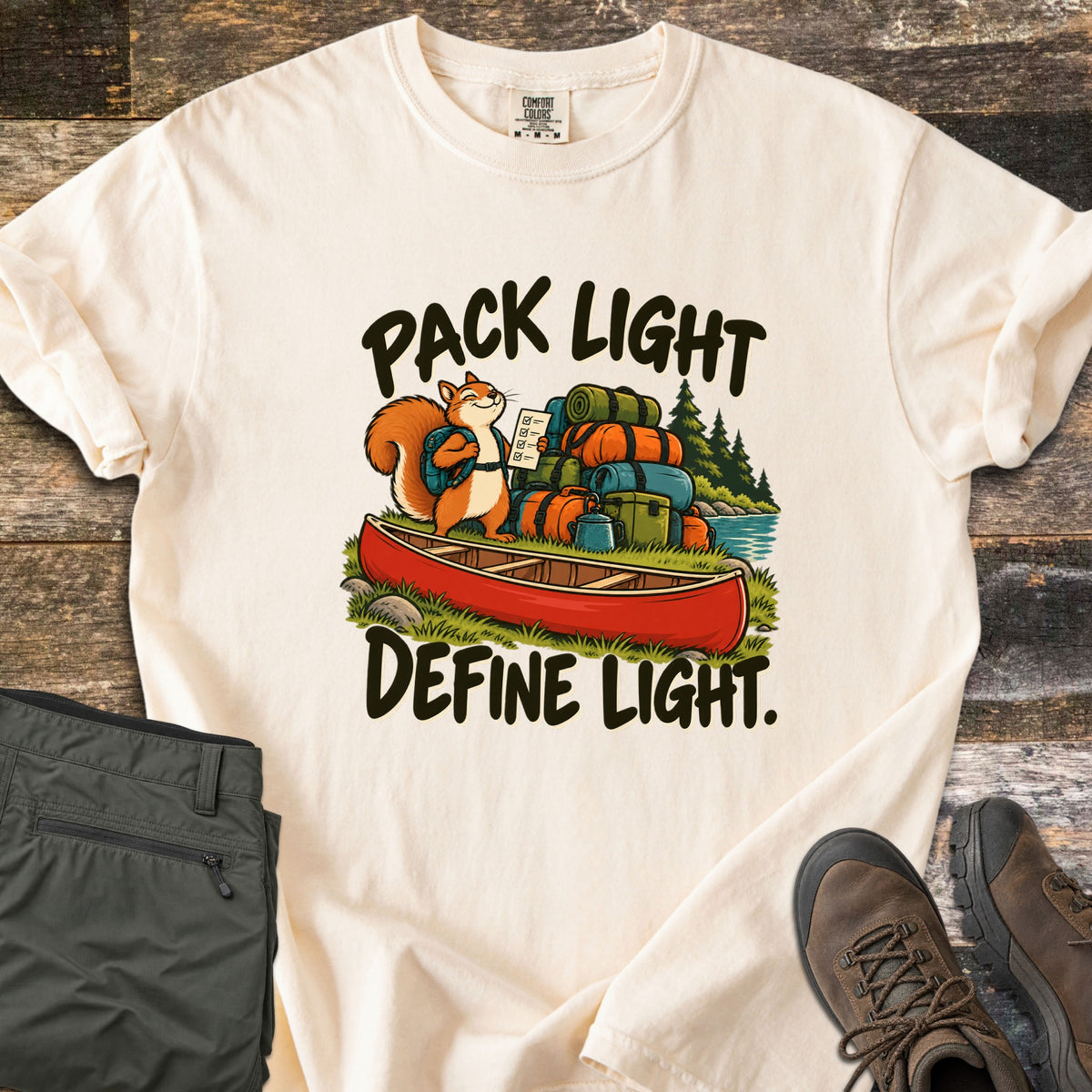 Pack Light Define Light Comfort Colors T-shirt in Ivory, laid flat. A smiling cartoon squirrel with a backpack holds a checklist beside a red canoe piled with gear bags, between the words Pack Light and Define Light. Olive pants and brown hiking boots sit on weathered wood.