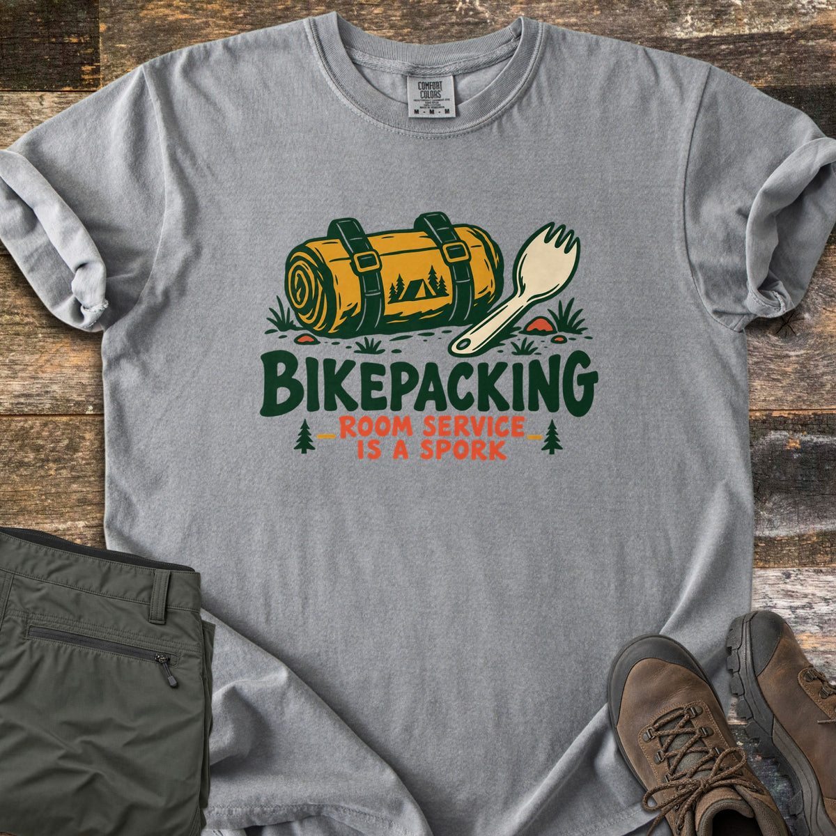 Bikepacking Room Service Is a Spork Comfort Colors T-shirt in Grey, laid flat. A strapped yellow bedroll showing a tent and pines lies next to a cream spork over the words Bikepacking Room Service Is a Spork. Sleeves are rolled on weathered wood with olive pants and brown boots.