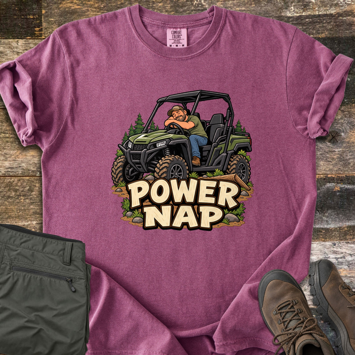 Power Nap Comfort Colors T-shirt in Berry, laid flat. A cartoon man in a cap naps with his head on the steering wheel of a green side-by-side UTV among pine trees, above the words Power Nap. Folded olive pants and brown hiking boots sit on a weathered wood surface.