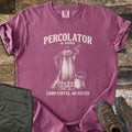 Percolator & Sons Comfort Colors T-shirt in Berry, laid flat. Cream ink drawing of a steaming percolator and tin mug on a campfire grate, under the words Percolator & Sons and above Camp Coffee, No Filter. Olive pants and brown hiking boots lie on weathered wood.