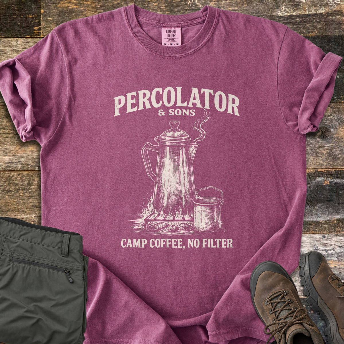 Percolator & Sons Comfort Colors T-shirt in Berry, laid flat. Cream ink drawing of a steaming percolator and tin mug on a campfire grate, under the words Percolator & Sons and above Camp Coffee, No Filter. Olive pants and brown hiking boots lie on weathered wood.