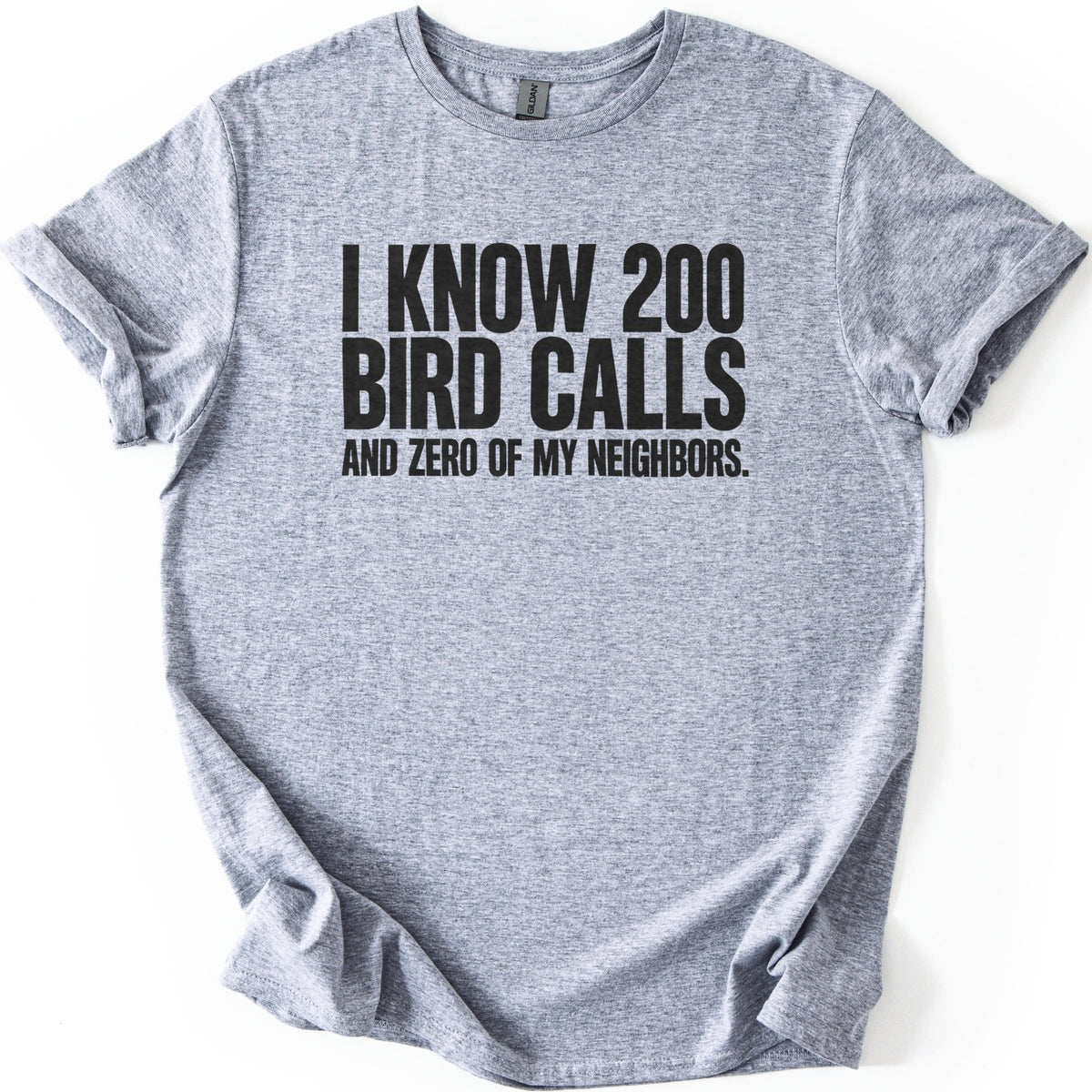 I Know 200 Bird Calls and Zero of My Neighbors Comfort Colors T-shirt in Sport Grey, laid flat. Bold black capital letters across the chest read I Know 200 Bird Calls and Zero of My Neighbors. The heathered shirt lies on a white background with rolled sleeves and a rumpled hem.