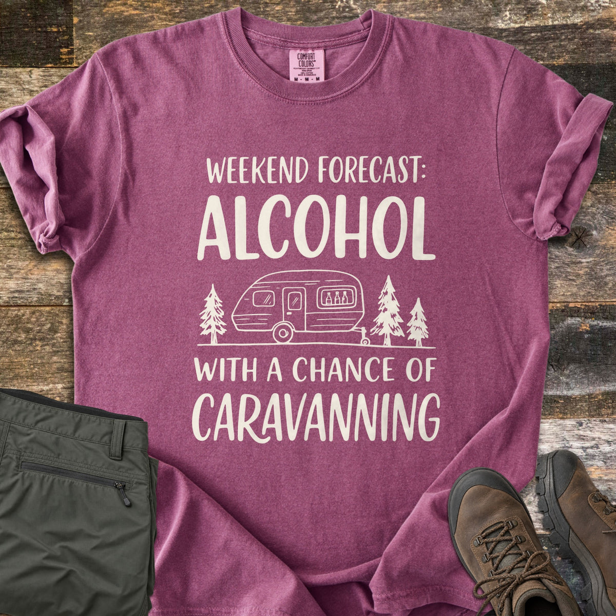 Alcohol With a Chance of Caravanning Comfort Colors T-shirt in Berry, laid flat. Cream print reads Weekend Forecast: Alcohol above a caravan with bottles in its window between pine trees, then With a Chance of Caravanning. Olive trousers and brown hiking boots sit on dark wood planks.