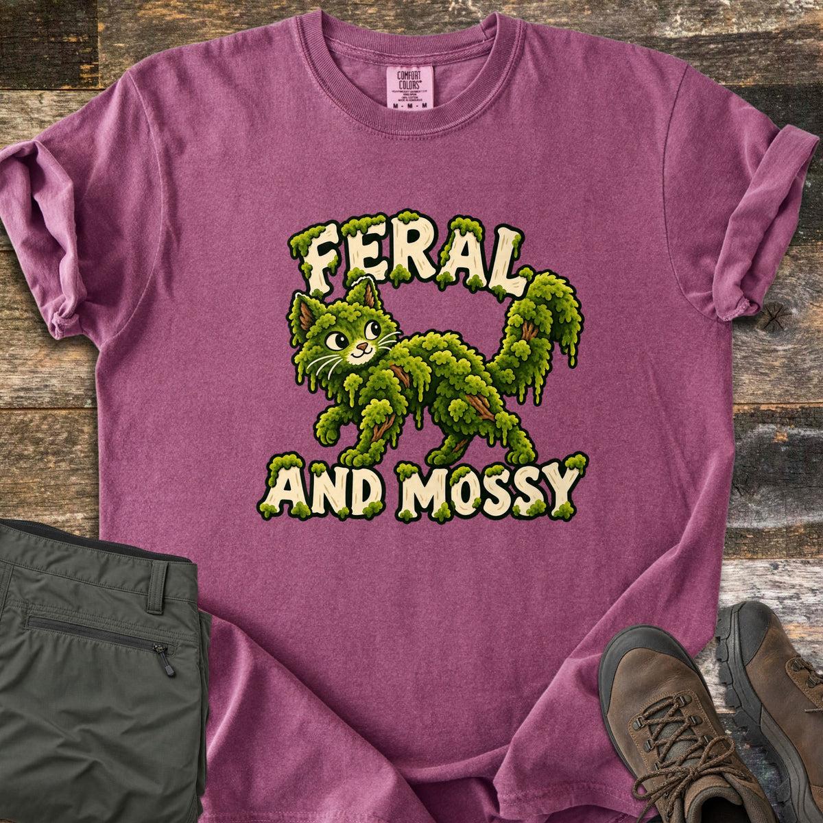 Feral and Mossy Comfort Colors T-shirt in Berry, laid flat. A cartoon cat made of green moss with bits of bark walks between the words Feral and Mossy in cream letters dripping with moss. Folded olive pants and brown hiking boots sit on a weathered wood floor.