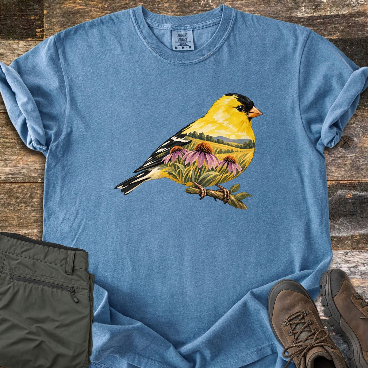 Goldfinch Meadow Comfort Colors T-shirt in Blue Jean, laid flat. A perched goldfinch whose body is filled with a meadow scene of purple coneflowers, grasses, hills and a pale sun. No text. Sleeves rolled, with olive cargo pants and brown hiking boots on old wood boards.