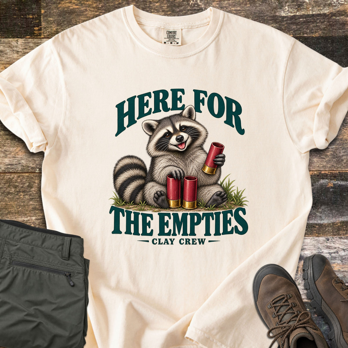 Here for the Empties Comfort Colors T-shirt in Ivory, laid flat. A smiling raccoon sits in grass holding red shotgun shells between the words Here for the Empties, with Clay Crew below. Olive cargo pants and brown hiking boots lie beside it on weathered wood.