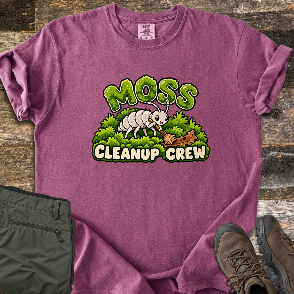 Moss Cleanup Crew Comfort Colors T-shirt in Berry, laid flat. A smiling white cartoon bug sweeps a brown leaf with a broom atop green moss, below the word MOSS in mossy letters and above CLEANUP CREW. It lies on weathered wood with folded olive pants and brown hiking boots.