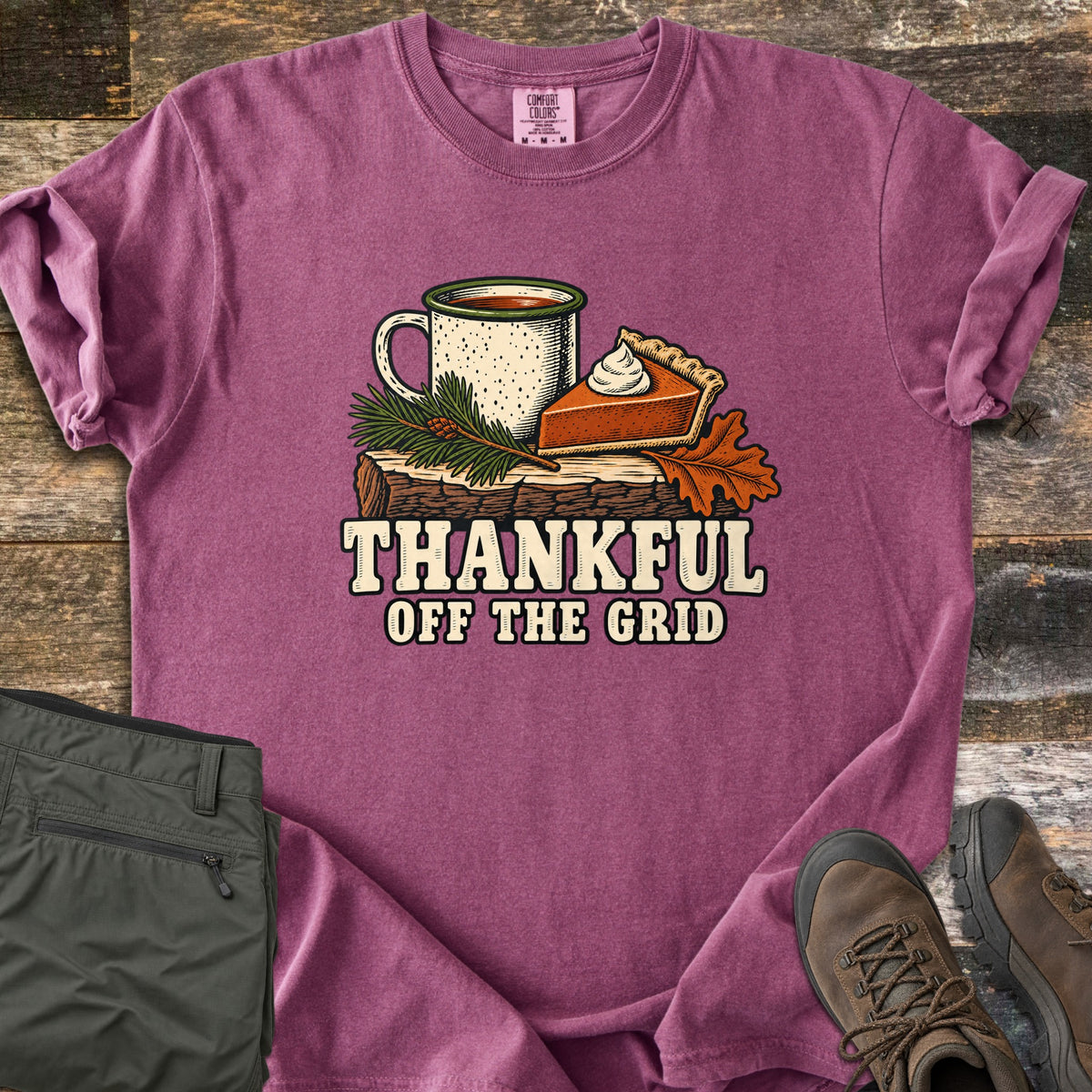 Thankful Off the Grid Comfort Colors T-shirt in Berry, laid flat. A speckled enamel camp mug and a slice of pumpkin pie with whipped cream sit on a log with a pine sprig and an oak leaf above the words Thankful Off the Grid. Olive hiking pants and brown boots lie on weathered wood.