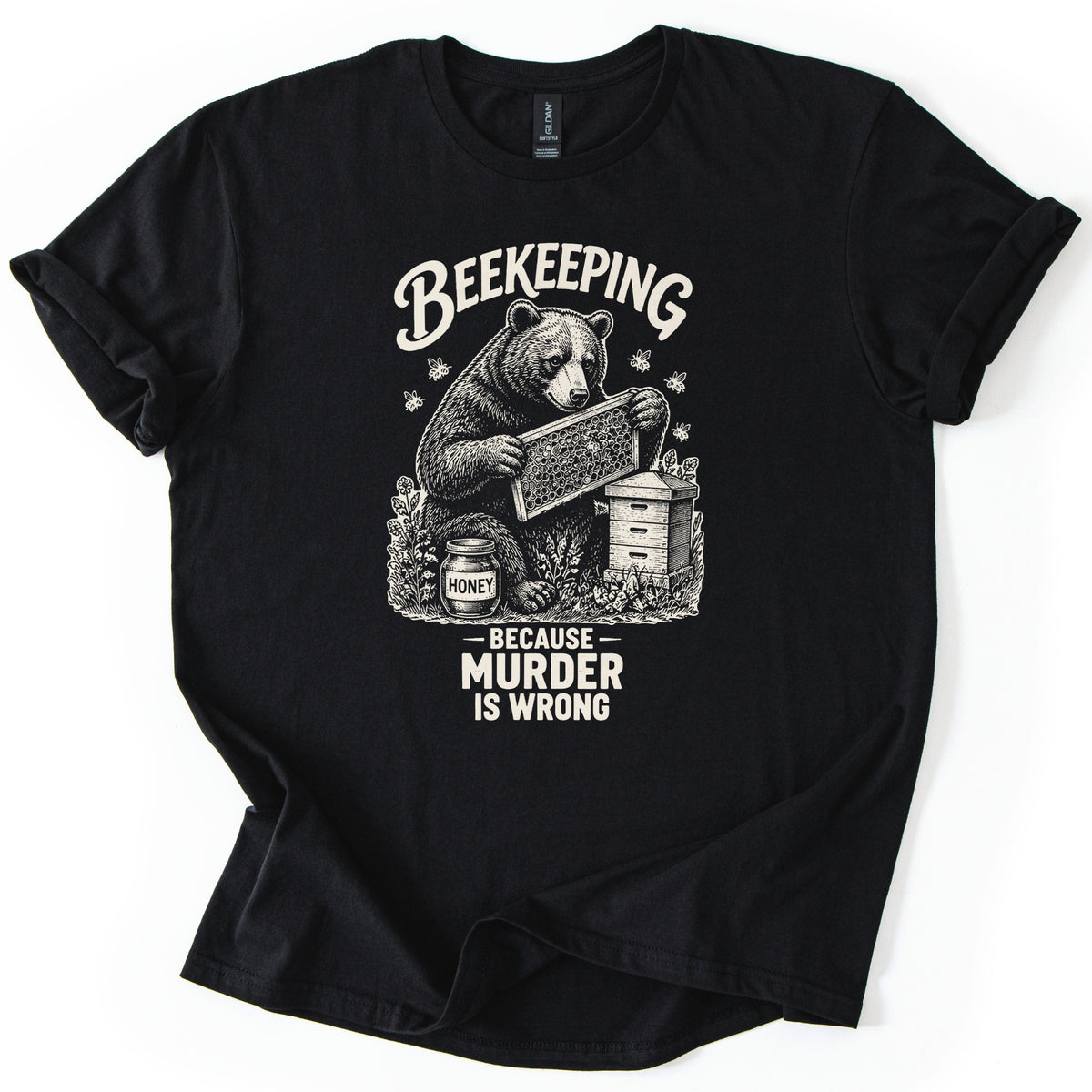 Beekeeping Murder Bear Comfort Colors T-shirt in Black, laid flat. A cream etched drawing of a bear holding a honeycomb frame by a beehive and a jar labeled Honey, with bees flying, under the word Beekeeping and above Because Murder Is Wrong. Shown on white with cuffed sleeves.