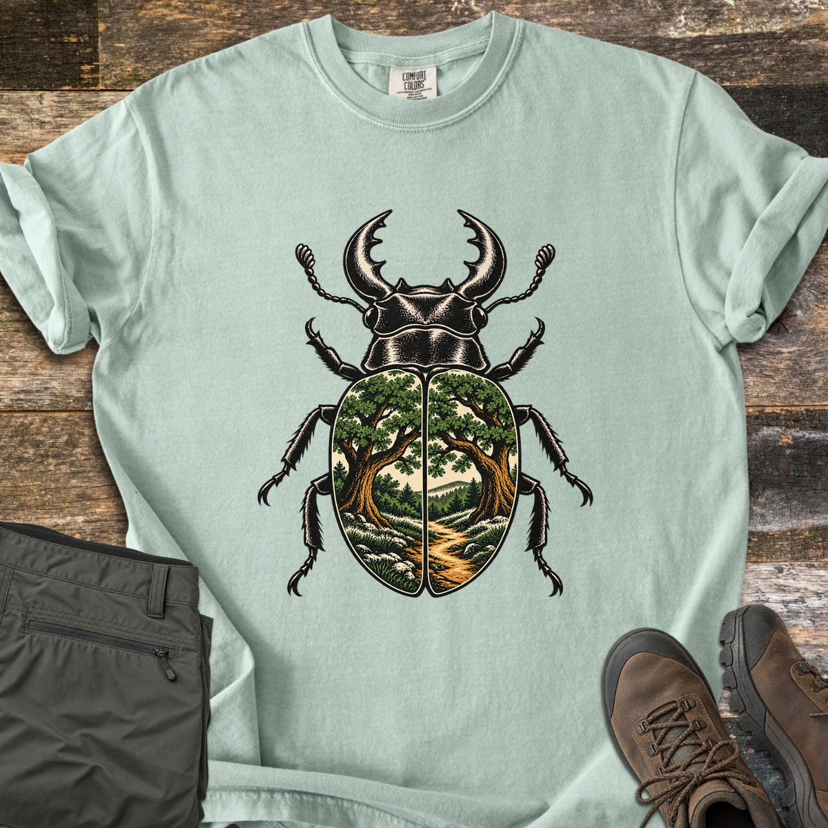 Beetle Forest Path Comfort Colors T-shirt in Bay, laid flat. A black stag beetle whose wing covers show a forest scene of two oak trees framing a dirt path toward distant hills. Folded olive hiking pants and brown hiking boots sit beside it on weathered wood planks.