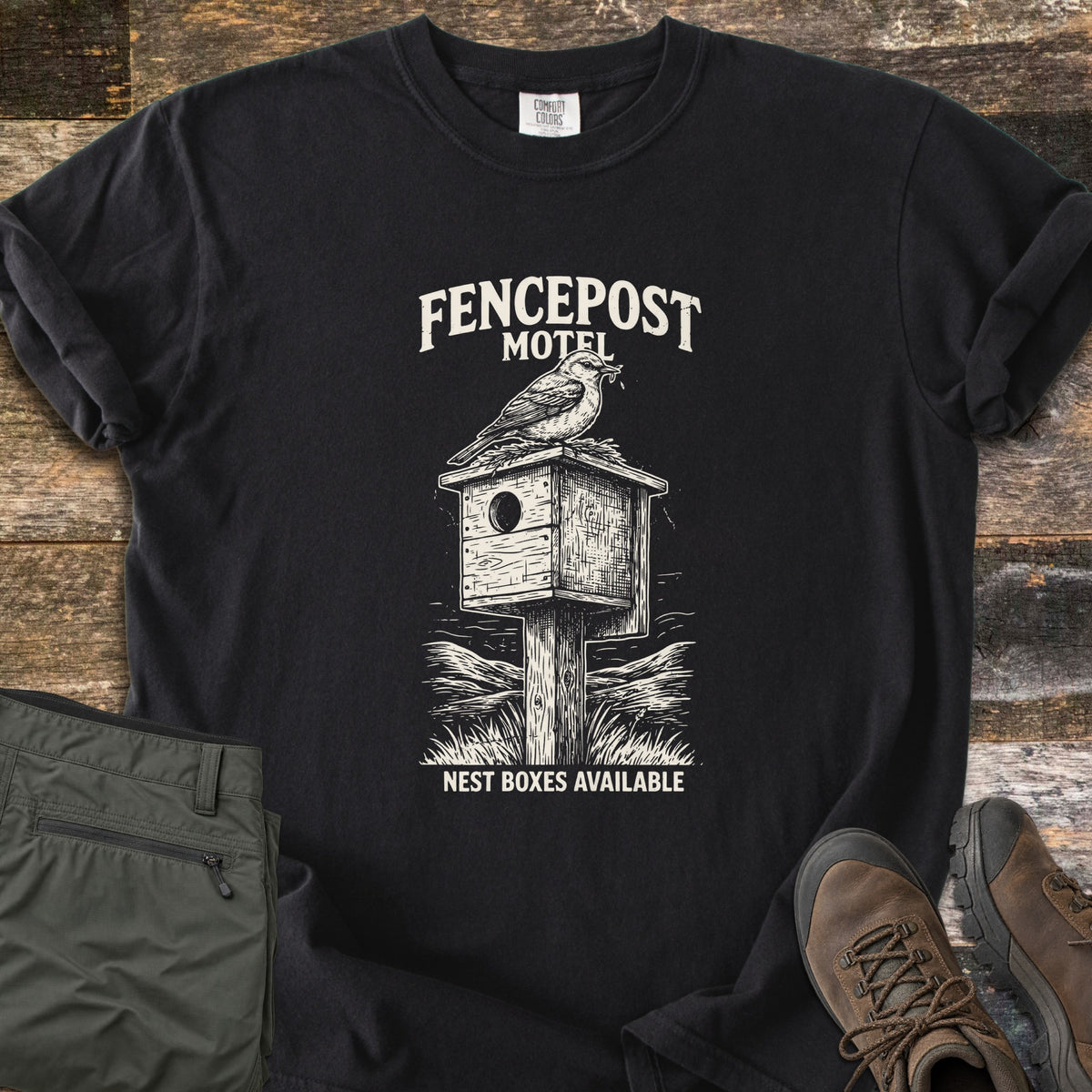 Fencepost Motel Comfort Colors T-shirt in Black, laid flat. A cream sketch of a bird perched on a wooden nest box atop a post in tall grass, with the words Fencepost Motel above and Nest Boxes Available below. Sleeves are rolled on a wood plank surface beside olive pants and brown hiking boots.