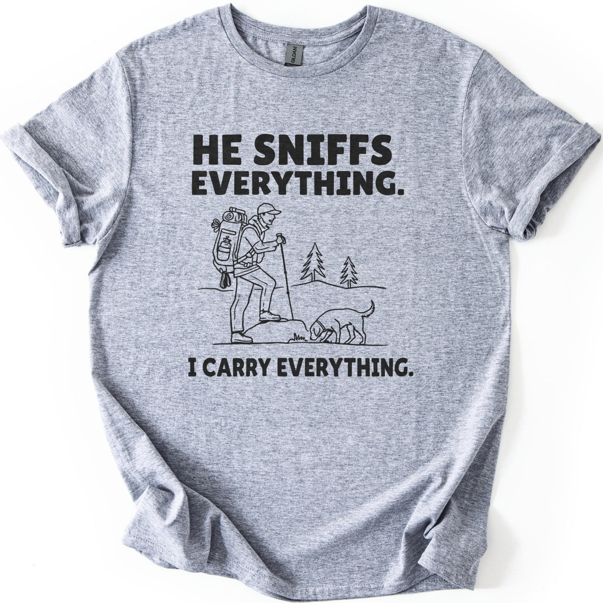 Trail Dog Pack Mule Comfort Colors T-shirt in Sport Grey, laid flat. Black line art of a hiker with a loaded backpack and trekking pole stepping onto a rock as a dog sniffs the ground, with pine trees behind, between the words He sniffs everything. and I carry everything. Sleeves rolled, on white.
