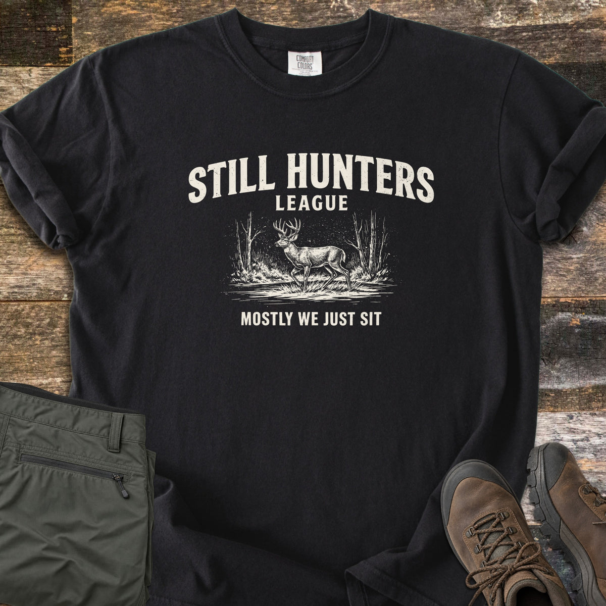 Still Hunters League Comfort Colors T-shirt in Black, laid flat. Cream print of the words Still Hunters League above a sketched buck deer standing in grass between bare trees, with Mostly We Just Sit below. Sleeves rolled, on weathered wood with olive pants and brown hiking boots.