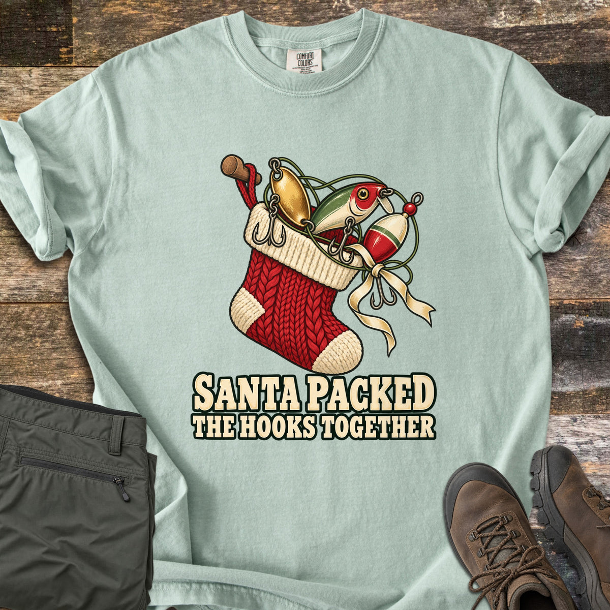 Santa Packed The Hooks Together Comfort Colors T-shirt in Bay, laid flat. A red knit Christmas stocking holds fishing lures, a bobber and a rod handle tied with a cream ribbon above the words Santa Packed The Hooks Together. Olive pants and brown boots sit on a wood floor.