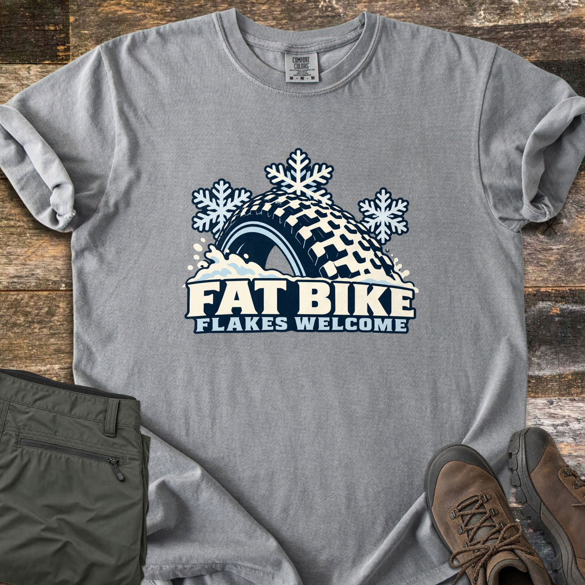 Fat Bike Flakes Welcome Comfort Colors T-shirt in Grey, laid flat. A fat bike tire half buried in snow with three snowflakes above it, over the words FAT BIKE FLAKES WELCOME. Sleeves are rolled, on a wood floor with folded olive pants and brown hiking boots.