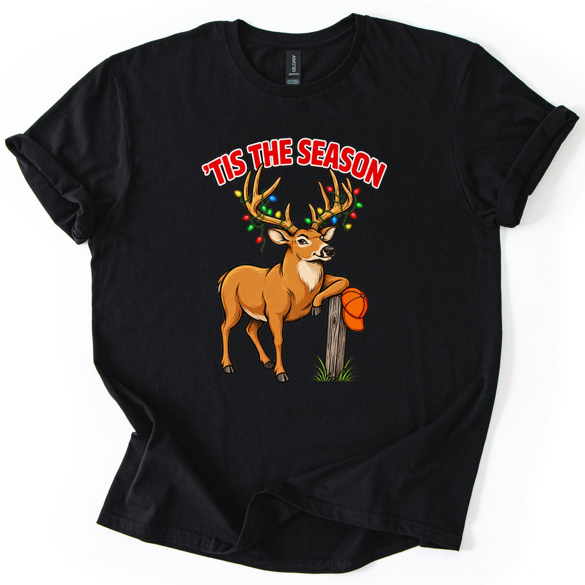 Buck Tis The Season Comfort Colors T-shirt in Black, laid flat. Red arched text reads 'TIS THE SEASON above a cartoon buck with colored Christmas lights strung on its antlers, leaning on a wooden post that holds an orange hunting cap. Sleeves rolled, on a white surface.