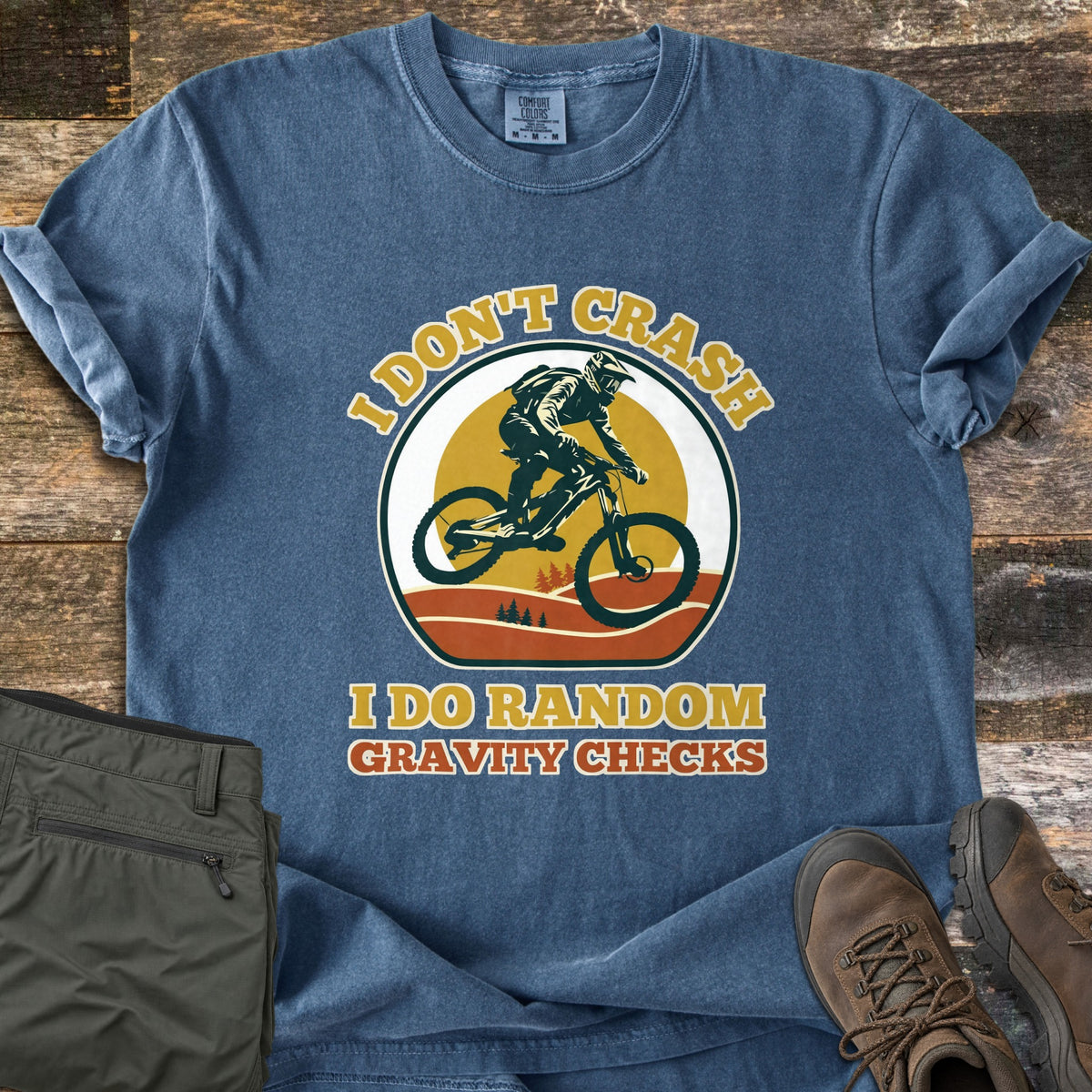 I Don't Crash I Do Random Gravity Checks Comfort Colors T-shirt in Denim, laid flat. A mountain biker in a full-face helmet rides over orange hills and pines before a yellow sun, between the words I Don't Crash and I Do Random Gravity Checks. Olive pants and brown hiking boots lie on weathered wood.