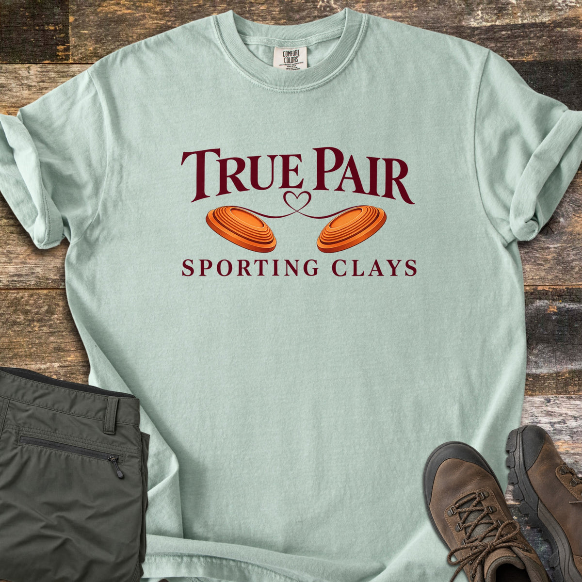 True Pair Comfort Colors T-shirt in Bay, laid flat. Dark red text reads True Pair above two orange clay targets joined by a looping heart line, with Sporting Clays below. Sleeves are rolled; olive pants and brown hiking boots sit on weathered wood.