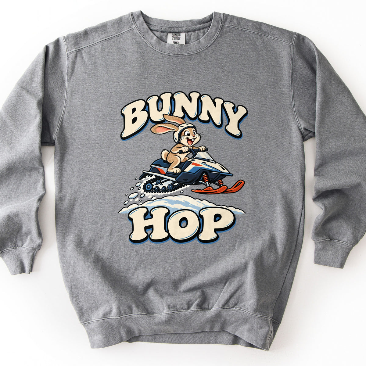 Bunny Hop Comfort Colors crewneck sweatshirt in Grey, laid flat. A grinning cartoon bunny in a white helmet rides a navy, white and orange snowmobile over a snowbank between the words Bunny Hop in cream bubble letters. The sweatshirt lies on a white surface.