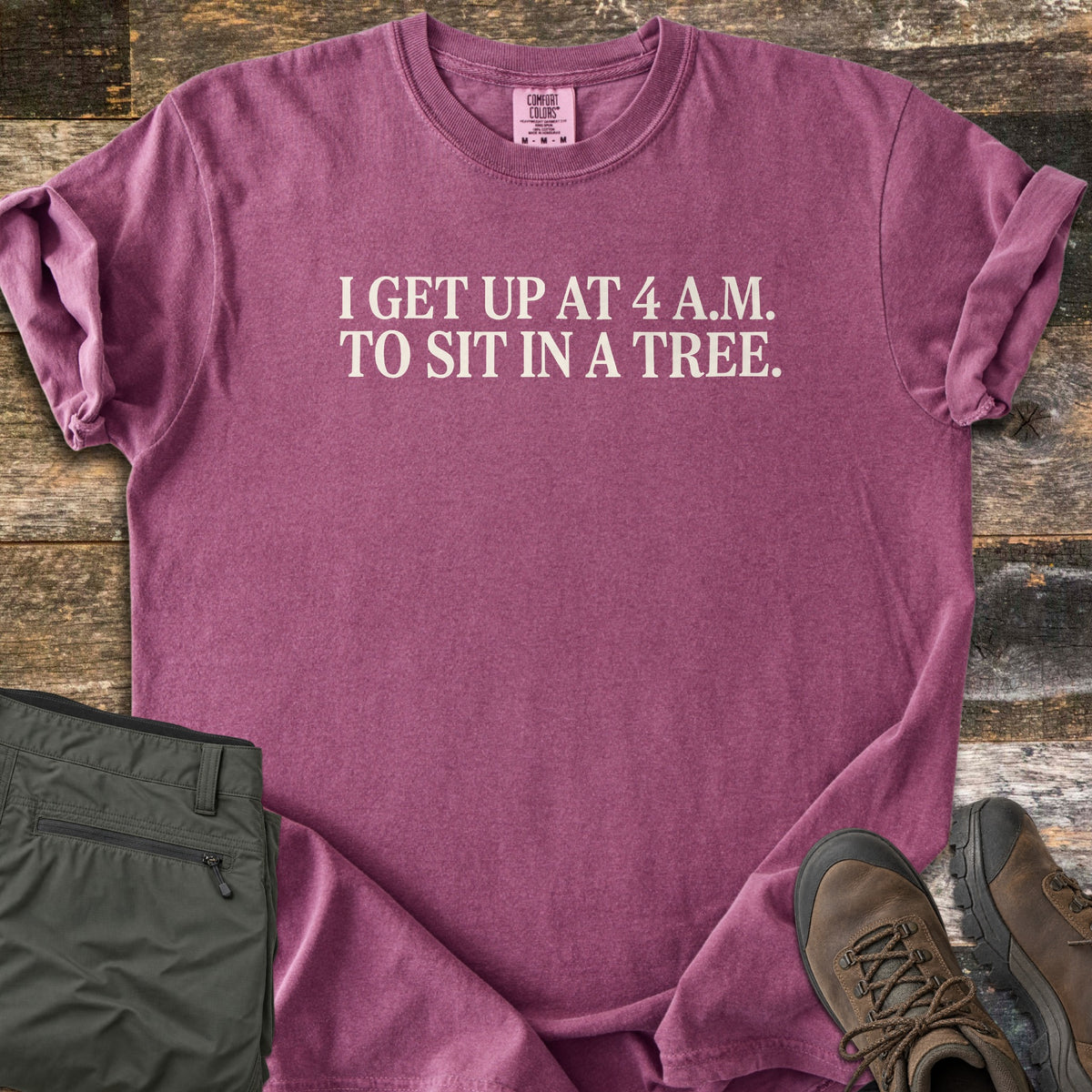Sit In A Tree Comfort Colors T-shirt in Berry, laid flat. Cream serif text reads I GET UP AT 4 A.M. TO SIT IN A TREE. across the chest. The shirt lies on weathered wood planks beside folded olive green cargo pants and brown leather hiking boots.