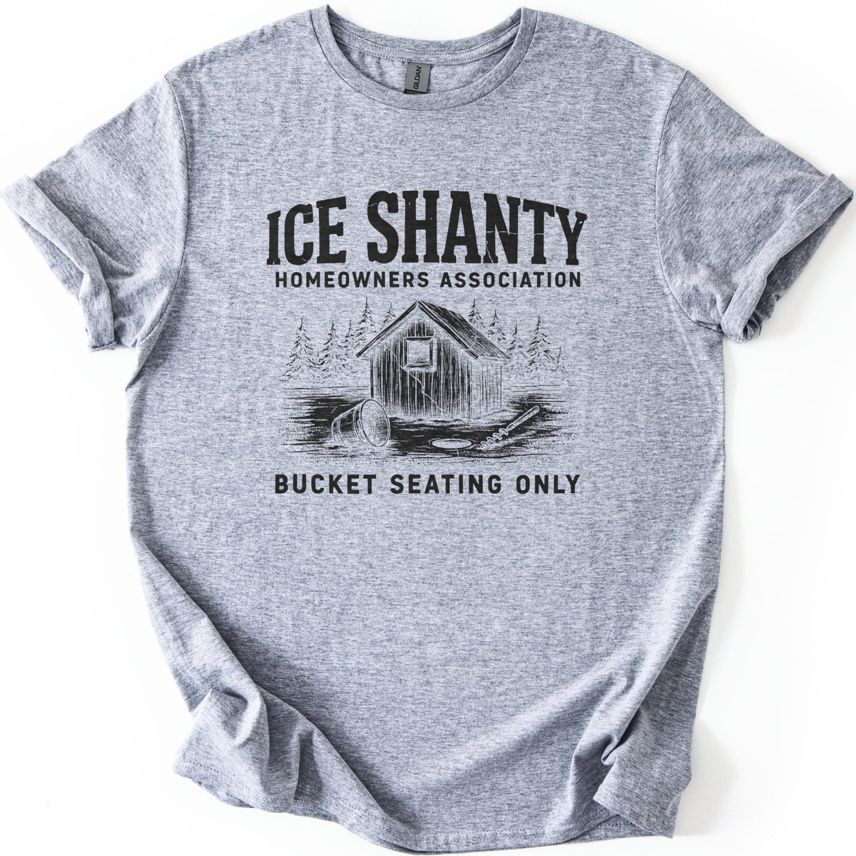 Ice Shanty Homeowners Association Comfort Colors T-shirt in Sport Grey, laid flat. Black print reads ICE SHANTY, HOMEOWNERS ASSOCIATION above a sketch of a wooden ice shack among pines with a tipped bucket, ice hole and auger, then BUCKET SEATING ONLY. Sleeves cuffed, on white.