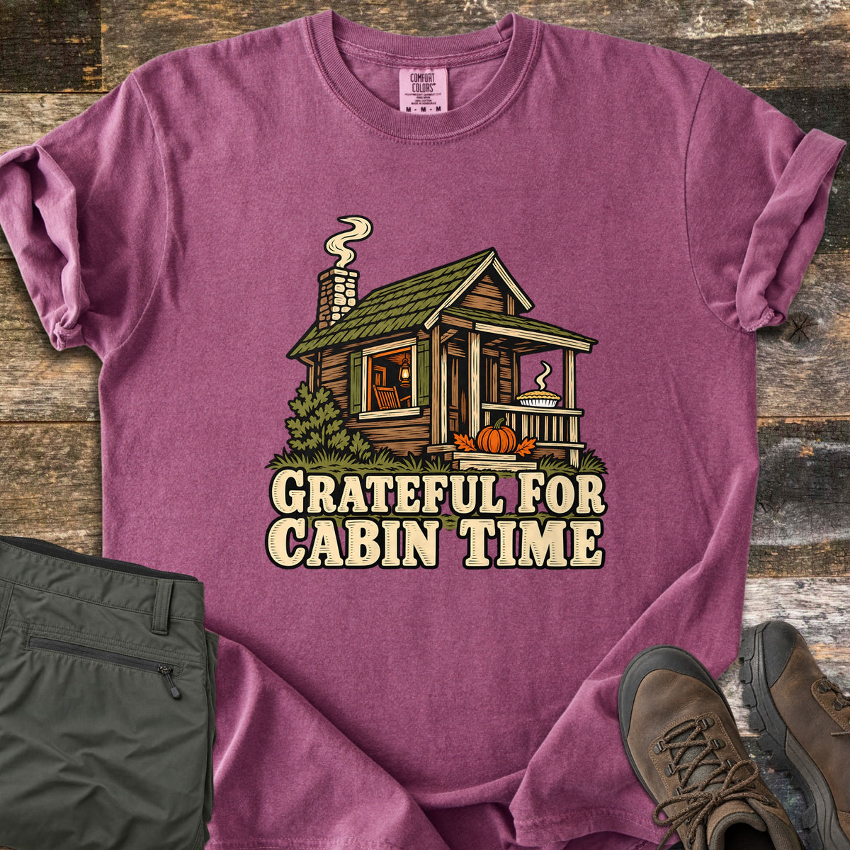 Grateful For Cabin Time Comfort Colors T-shirt in Berry, laid flat. Print of a log cabin with a green roof, smoking chimney, a pie on the porch rail and a pumpkin on the steps, above the words Grateful For Cabin Time. Green pants and brown hiking boots lie on weathered wood.