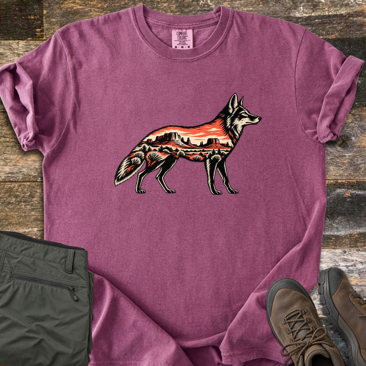Desert Mesa Coyote Comfort Colors T-shirt in Berry, laid flat. A side-view coyote silhouette filled with a red desert mesa landscape, sunset sky and shrubs, with no words. Sleeves rolled on rustic wood, beside folded olive hiking pants and brown hiking boots.