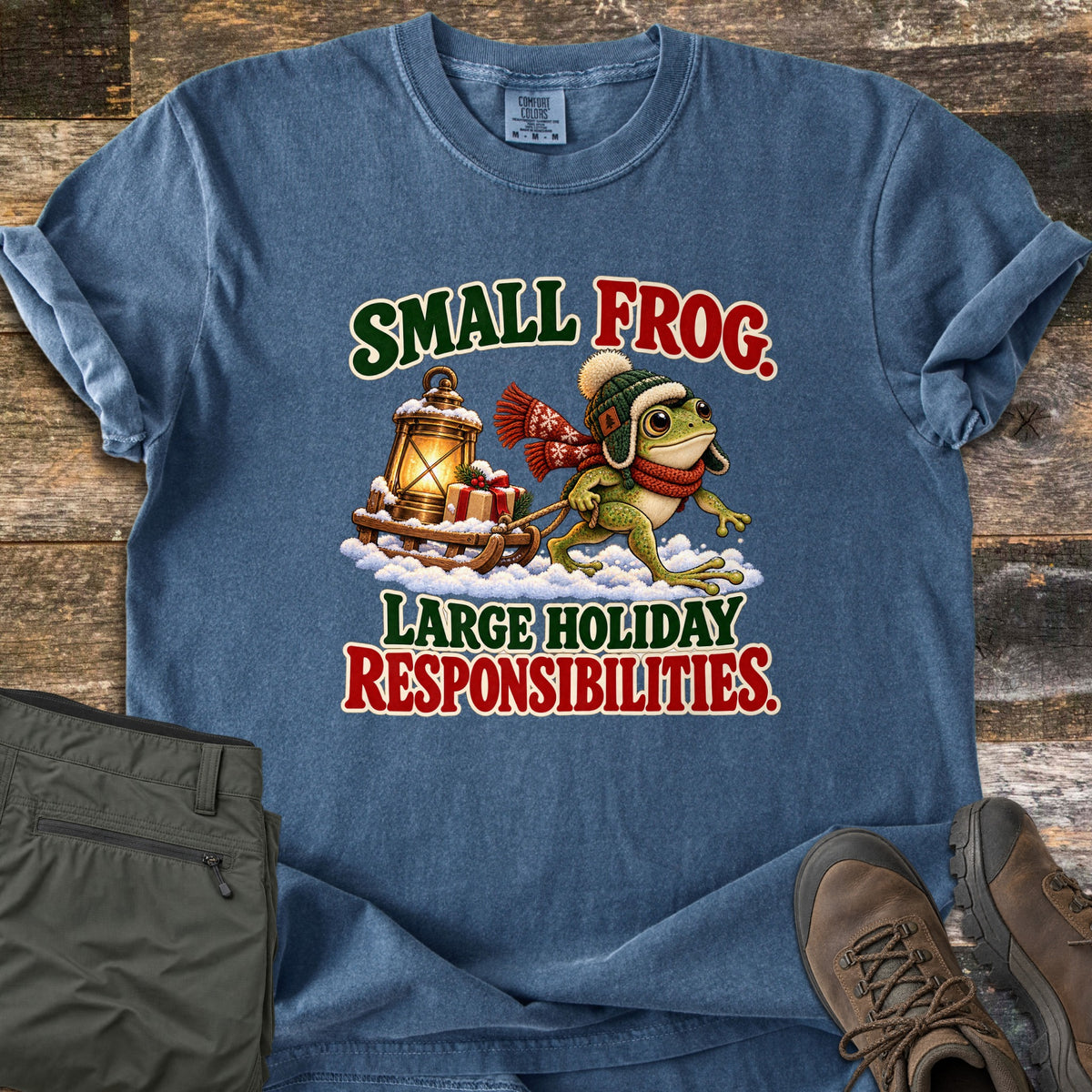 Small Frog. Large Holiday Responsibilities Comfort Colors T-shirt in Denim, laid flat. A frog in a green knit hat and red scarf pulls a wooden sled with a lantern and gift through snow, between the words Small Frog. Large Holiday Responsibilities. Olive pants and brown boots sit on weathered wood.