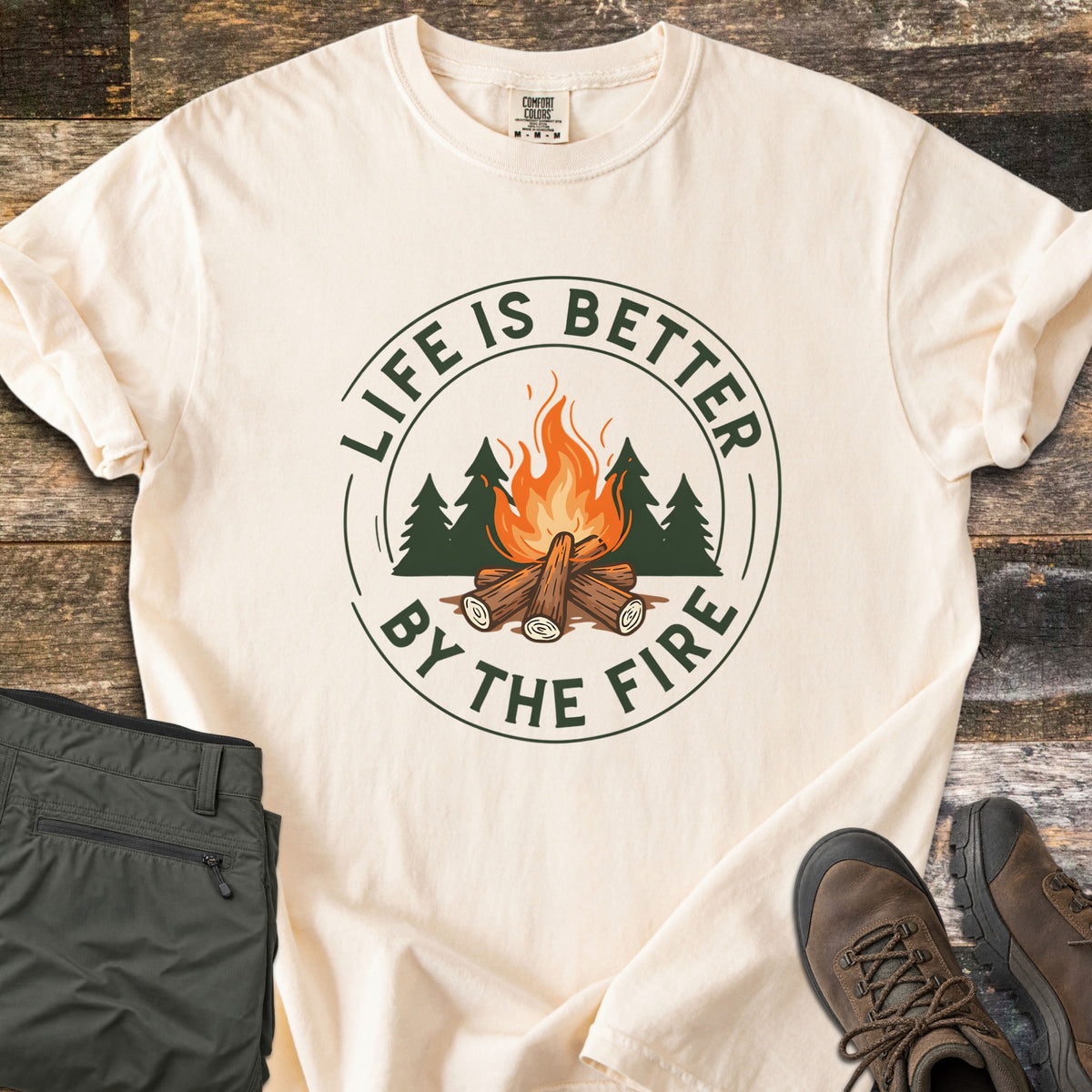 Life Is Better By The Fire Ring Comfort Colors T-shirt in Ivory, laid flat. A round badge in dark green and orange shows a log campfire in front of pine trees, ringed by the words Life Is Better By The Fire. Olive hiking pants and brown hiking boots lie beside it on weathered wood planks.