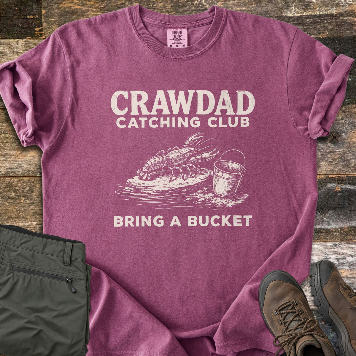 Crawdad Catching Club Comfort Colors T-shirt in Berry, laid flat. Cream print reads CRAWDAD CATCHING CLUB above a sketch of a crawdad on a rock beside a bucket, with BRING A BUCKET below. Folded olive pants and brown hiking boots lie beside it on weathered wood.