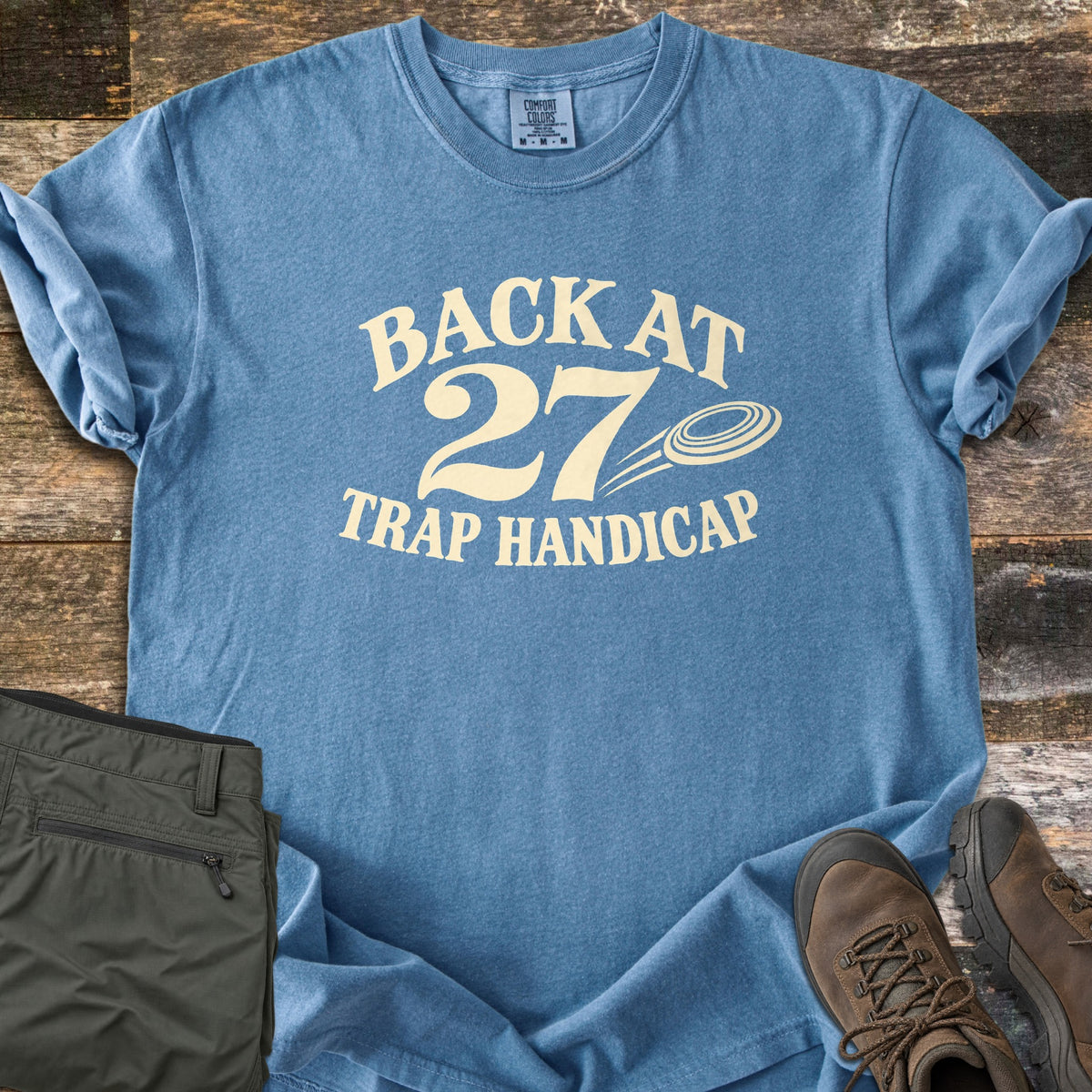 Back at 27 Trap Handicap Comfort Colors T-shirt in Blue Jean, laid flat. Cream print reads Back at 27 in an arch above Trap Handicap, with a clay target flying beside the 27. Sleeves rolled on a rustic wood surface, with olive cargo pants and brown hiking boots.