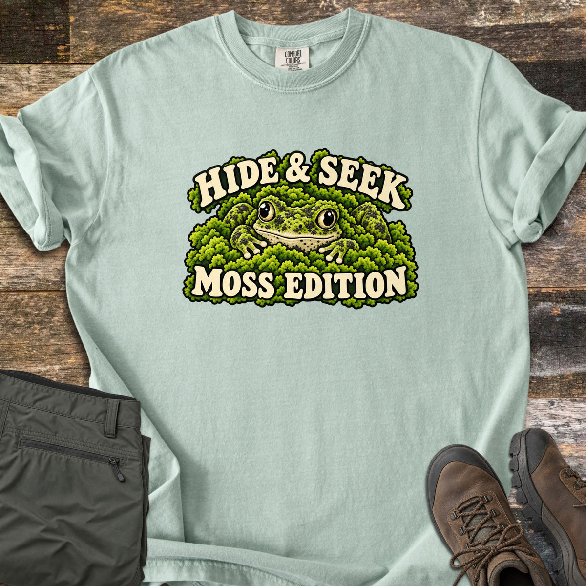 Hide & Seek Moss Edition Comfort Colors T-shirt in Bay, laid flat. A wide-eyed frog peeks out from a mound of green moss between the words Hide & Seek and Moss Edition. Olive cargo pants and brown hiking boots lie beside it on weathered wood planks.